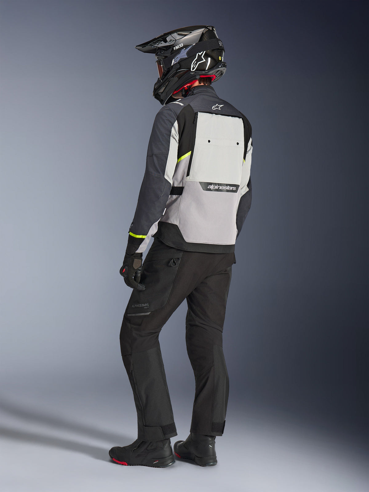 Veste Alpinestars Cusco Drystar®, coloris gris glacé, gris moyen et gris foncé, vue arrière portée par un pilote, accents jaune fluo, large panneau de ventilation au dos avec logo, construction textile durable, tenue de touring complète avec pantalon noir et casque d'aventure.