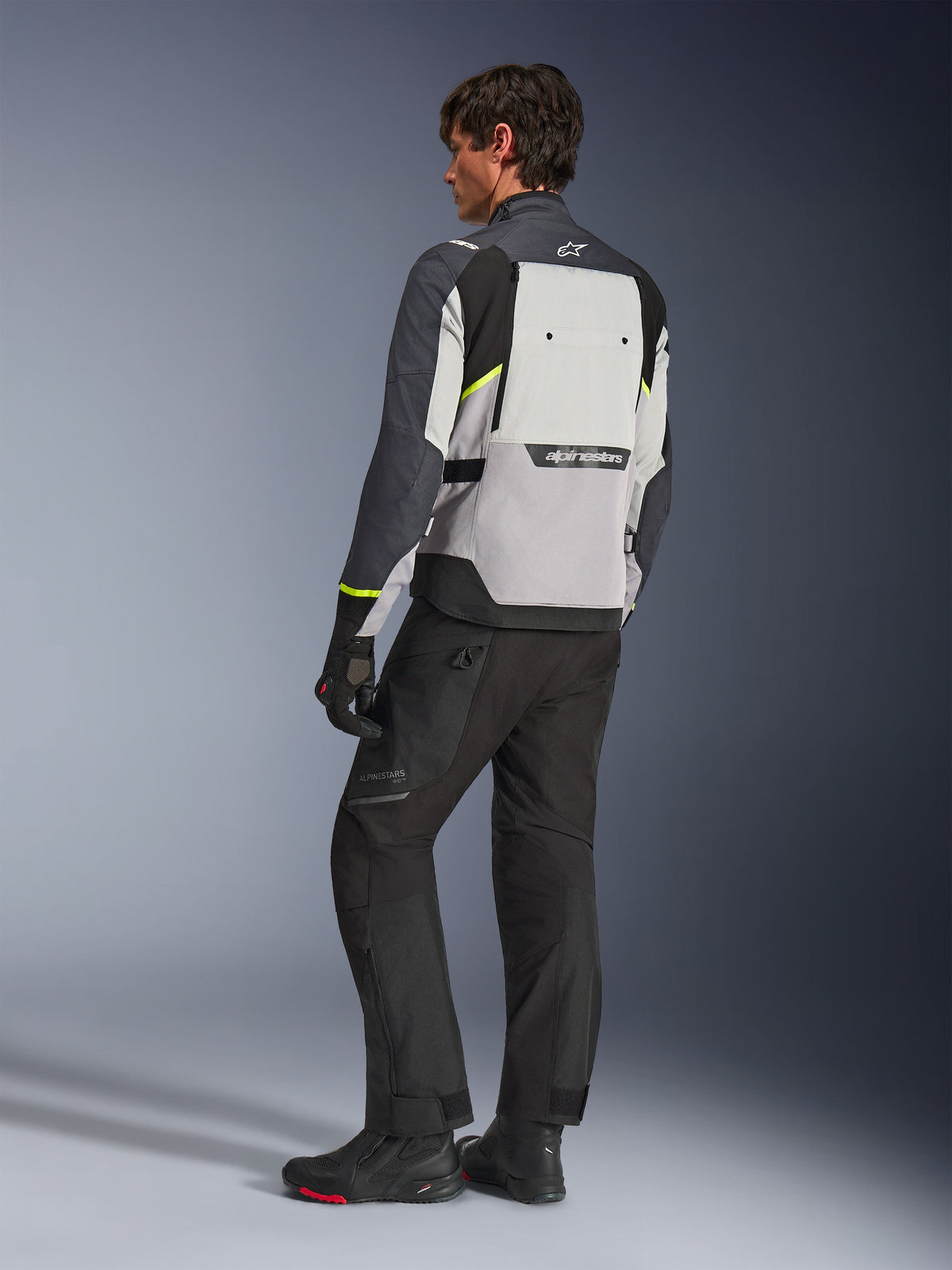 Veste Alpinestars Cusco Drystar®, coloris Ice Gray, Mid Gray et Deep Gray, vue de dos sur mannequin, veste de touring aventure avec accents jaune fluo, portée avec un pantalon et des bottes de moto noirs, dotée d'un large panneau de ventilation dorsal et du logo Alpinestars.