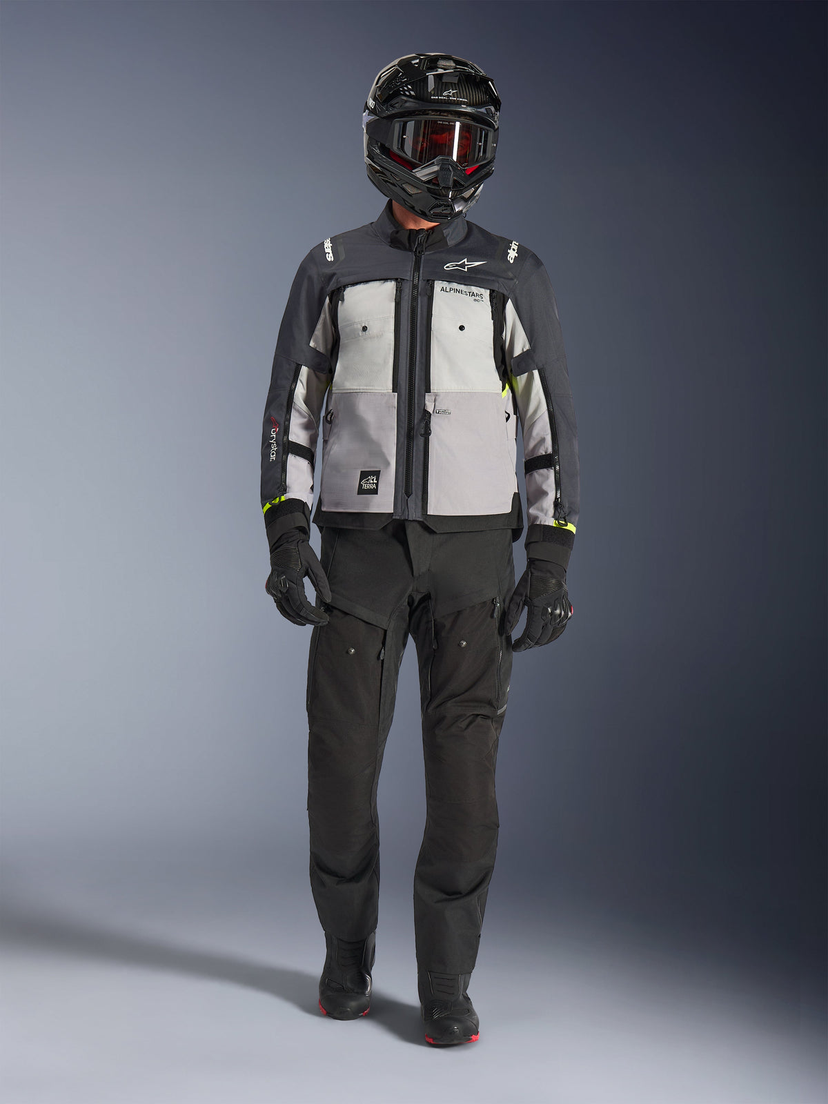 Veste Alpinestars Cusco Drystar®, Gris Glace, Gris Moyen et Gris Foncé, veste de moto d'aventure portée avec un pantalon de moto noir, des gants noirs et un casque noir avec lunettes, dotée de panneaux gris contrastés et de détails techniques pour la route et le touring d'aventure.