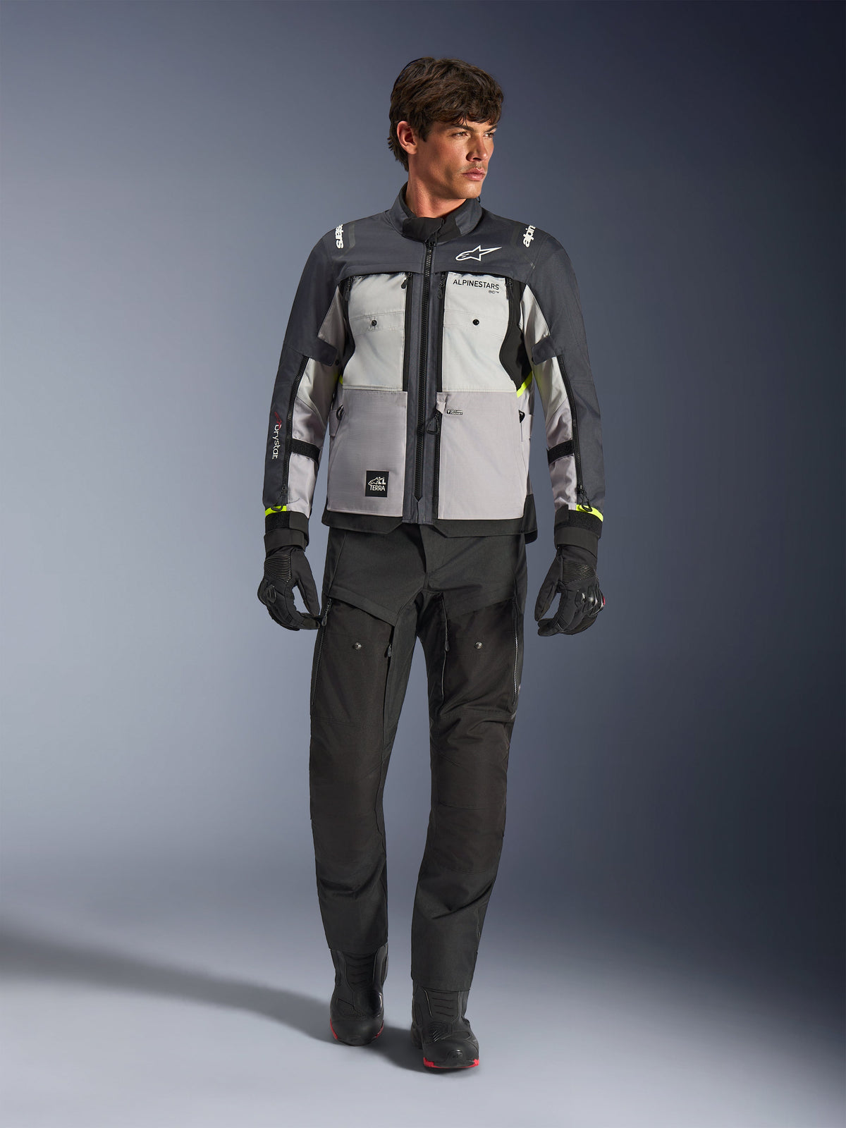 Veste Alpinestars Cusco Drystar® en Ice Gray, Mid Gray et Deep Gray, portée avec un pantalon et des gants de moto adventure noirs, veste de touring adventure en textile avec panneaux gris multi-tons et accents jaune fluo, photo de studio d'un mannequin homme.