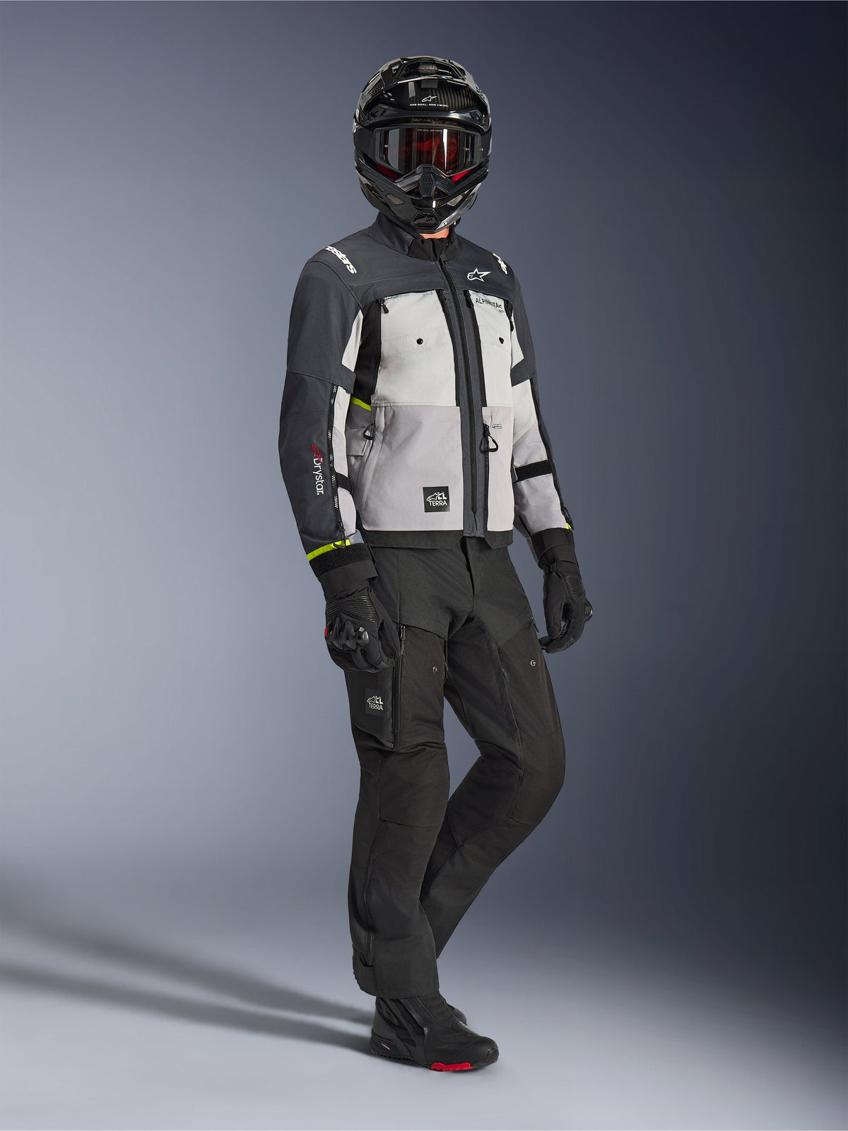 Veste Alpinestars Cusco Drystar® en Ice Gray Mid Gray Deep Gray, vue en pied d'un motard portant un équipement d'aventure complet comprenant un casque noir avec des lunettes rouges, un pantalon noir assorti et des gants, veste de moto en textile avec panneaux de ventilation directe et membrane imperméable.