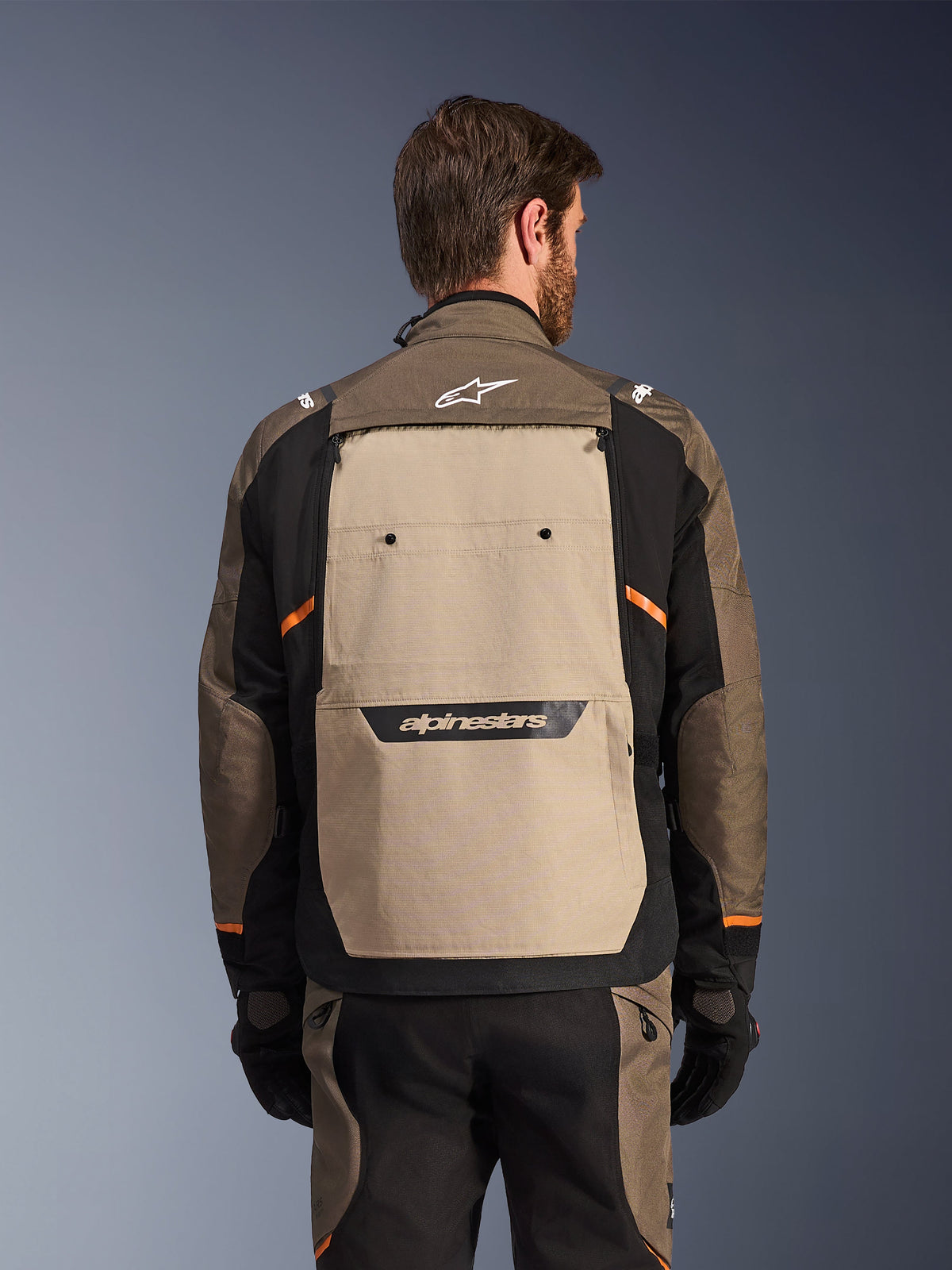 Veste Alpinestars Cusco Drystar®, Walnut Aluminum Black, vue arrière, présentant un large panneau dorsal beige avec logo noir et accents orange, portée avec un pantalon et des gants assortis, conçue pour le moto-tourisme d'aventure.