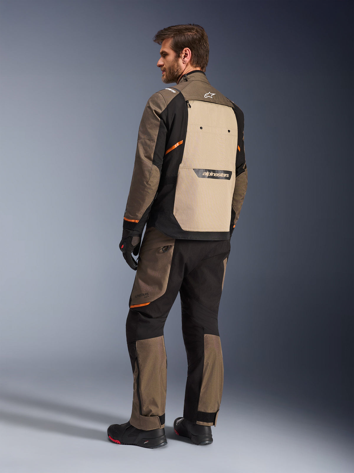 Veste Alpinestars Cusco Drystar® en Walnut Aluminum Black, vue arrière montrant le panneau dorsal beige et des touches d'orange, portée avec un pantalon de moto assorti, des gants et des bottes noirs, équipement professionnel d'aventure touring