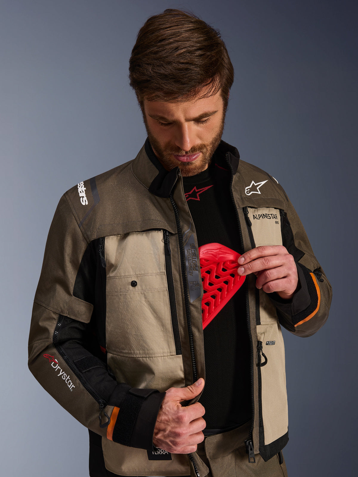 Veste Alpinestars Cusco Drystar®, Walnut Aluminum Black, portée par un homme montrant la poche pour protection pectorale avec un insert de protection rouge, veste de moto en textile avec ventilations zippées et logos pour l'aventure touring.