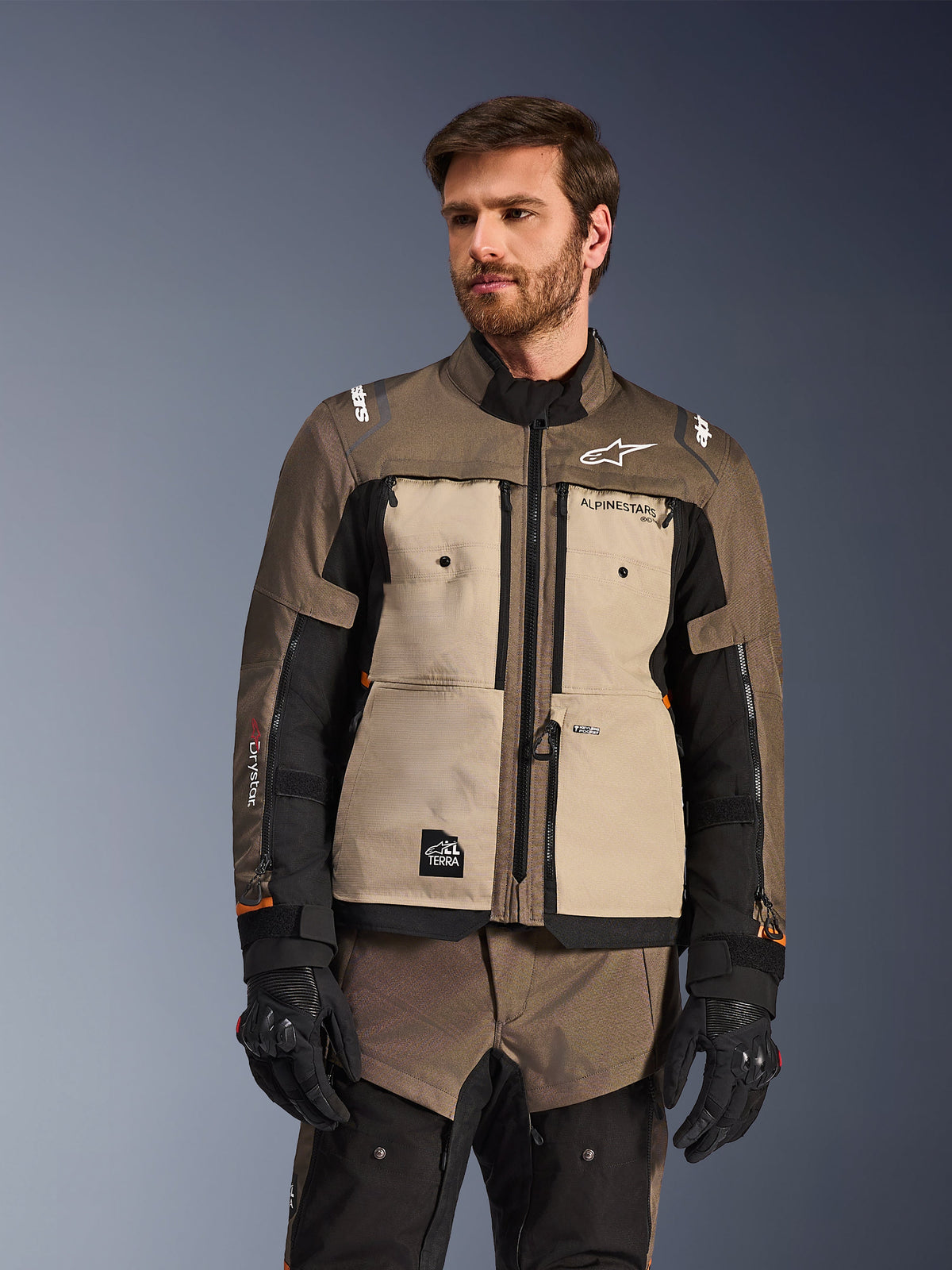 Veste Alpinestars Cusco Drystar®, veste de moto aventure, Walnut Aluminum Black, portée par un mannequin avec des gants noirs, avec des empiècements de poitrine beiges et des manches noires arborant le logo Alpinestars et l'écusson All Terra, équipement de touring de protection toutes saisons