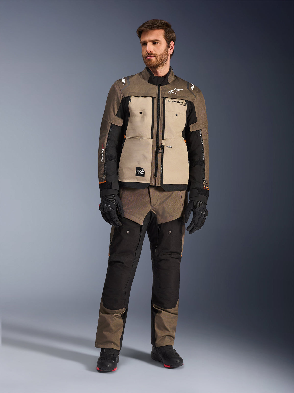 Veste Alpinestars Cusco Drystar® en Walnut Aluminum Black, portée par un motard avec un pantalon d'aventure touring assorti, des gants et des bottes noirs, dotée de panneaux de poitrine beiges, de manches noires avec de subtils accents orange et de fermetures éclair de ventilation fonctionnelles pour la conduite moto sur route.