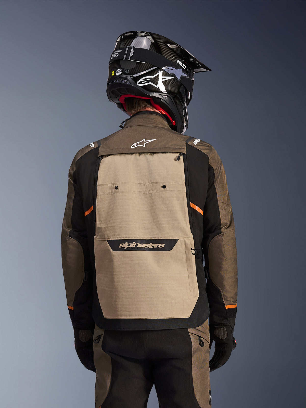 Vue arrière de la veste Alpinestars Cusco Drystar® en Walnut Aluminum Black, veste de moto d'aventure touring dotée d'un large empiècement dorsal beige avec logo Alpinestars, manches noires avec accents orange, présentée avec un casque noir style carbone.