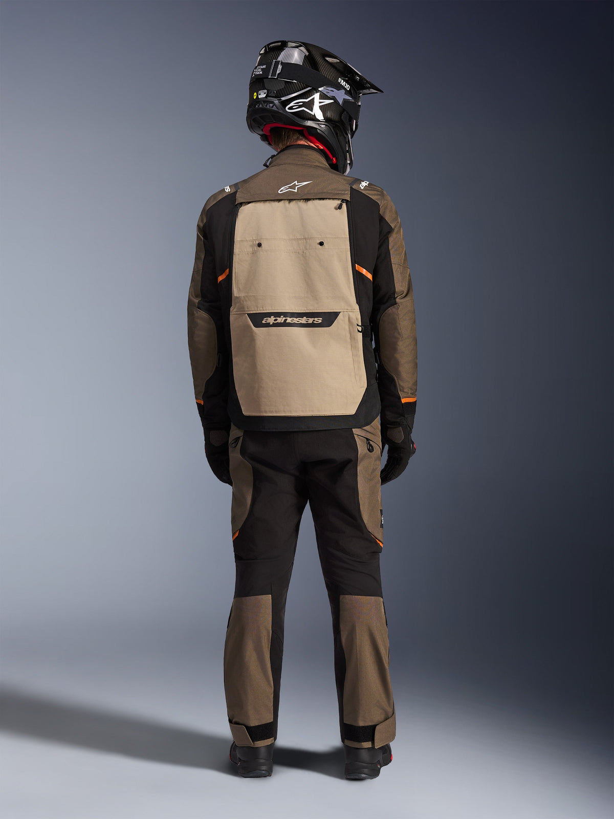 Motard de dos portant la veste Alpinestars Cusco Drystar® en Walnut Aluminum Black, avec une grande poche arrière couleur aluminium et des accents orange, pantalon d'aventure assorti, casque tout-terrain noir et gants, équipement moto en textile durable pour le touring.