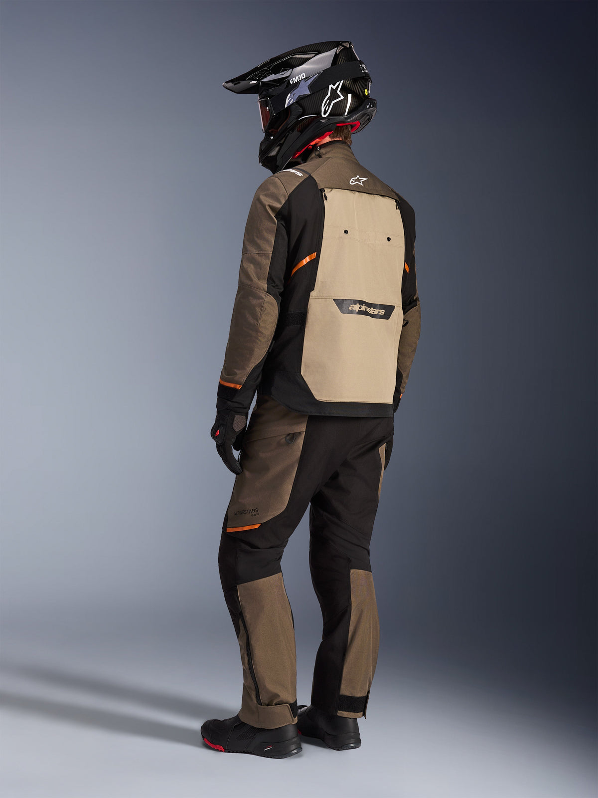 Veste Alpinestars Cusco Drystar®, veste d'aventure touring, Walnut Aluminum Black, vue arrière montrant le panneau arrière beige et des accents orange, portée avec un pantalon assorti et un casque noir en fibre de carbone.