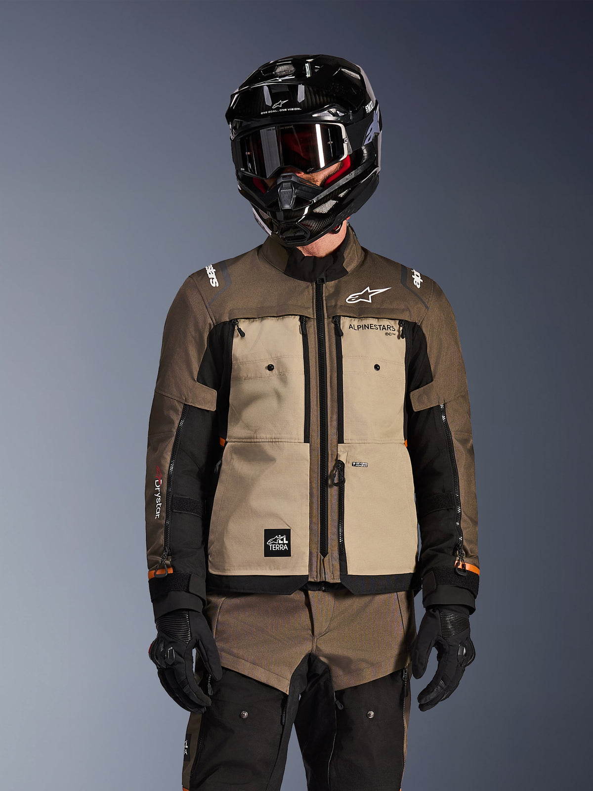 Motard portant la veste Alpinestars Cusco Drystar en Walnut Aluminum Black, tenue de moto adventure touring comprenant un casque noir avec lunettes et des gants noirs, vue de face montrant la construction textile technique, les poches et le logo.