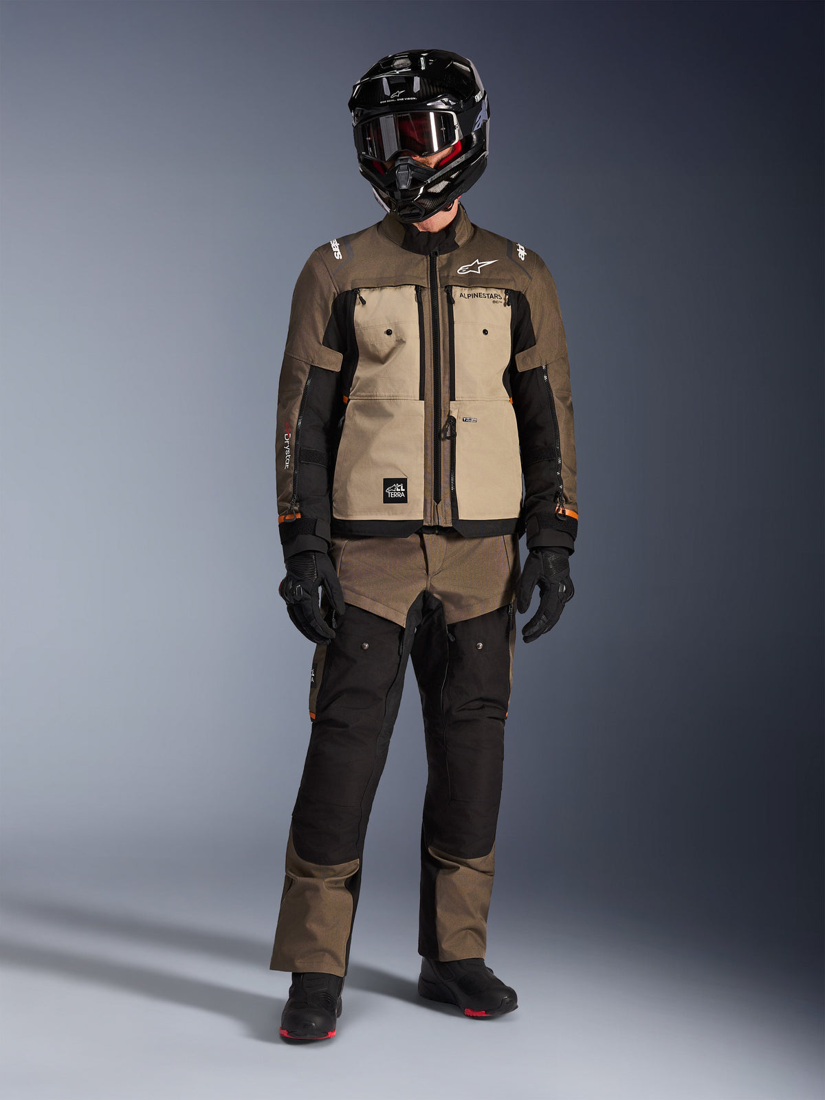 Veste Alpinestars Cusco Drystar®, Walnut Aluminum Black, veste de moto d'aventure touring portée avec un pantalon assorti et un casque noir, dotée de panneaux de poitrine beiges et de manches en ripstop marron, équipement technique de conduite toutes saisons