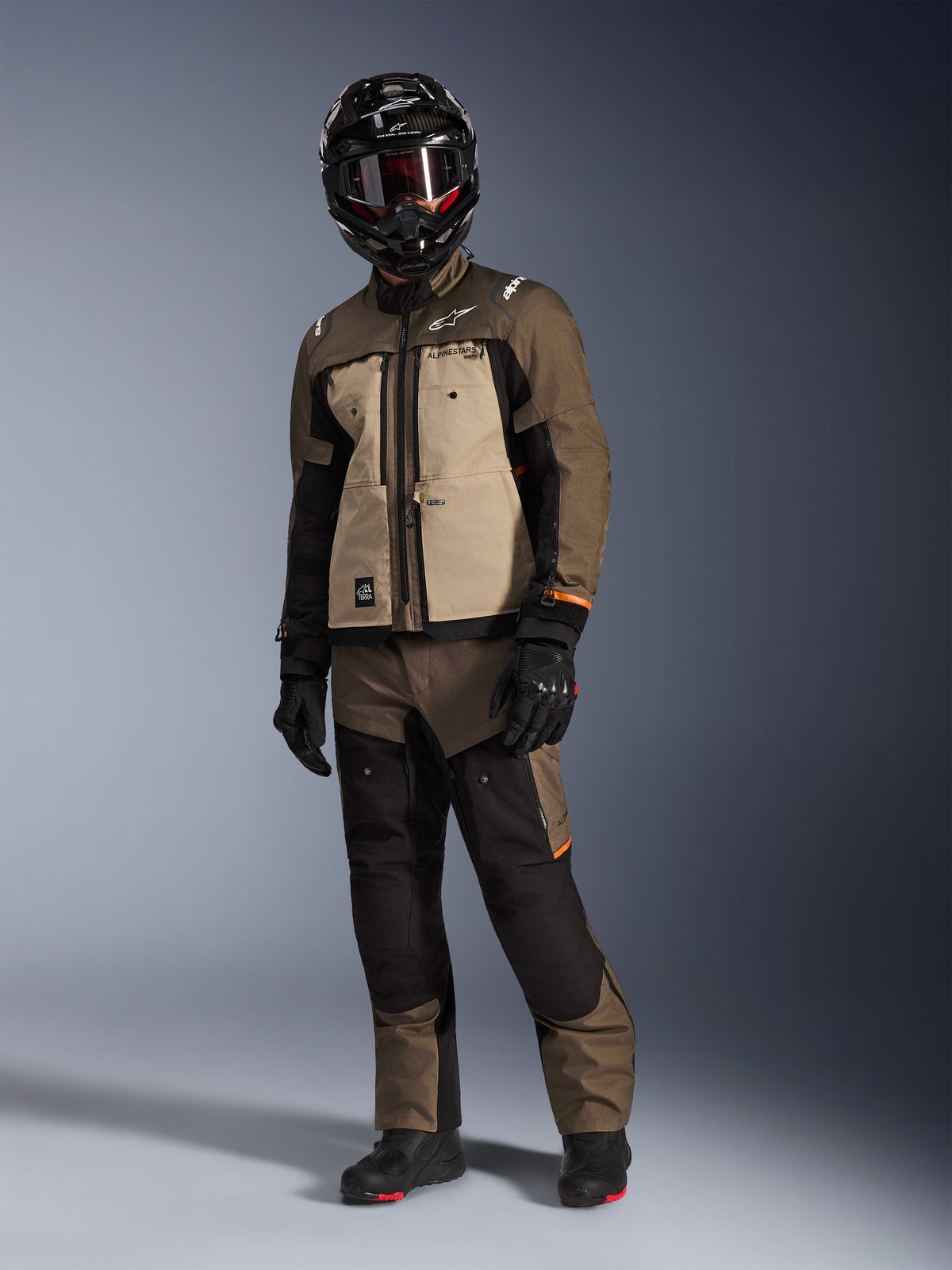 Veste Alpinestars Cusco Drystar®, Walnut Aluminum Black, portée par un motard en équipement d'aventure touring complet, avec empiècements beige et marron et détails noirs, pantalon assorti, casque intégral noir avec lunettes et bottes noires, fond de studio.
