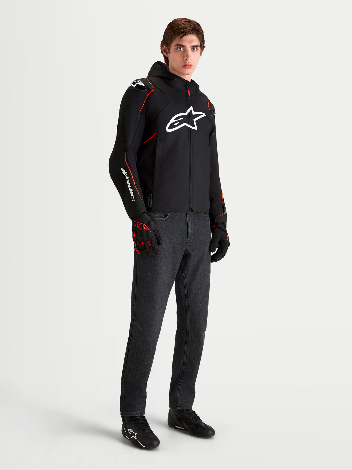 Veste imperméable Zaca V 2 d'Alpinestars, veste de moto sport pour homme noire et rouge avec capuche et logo blanc sur la poitrine, portée par un modèle masculin avec des gants de moto assortis et un jean foncé