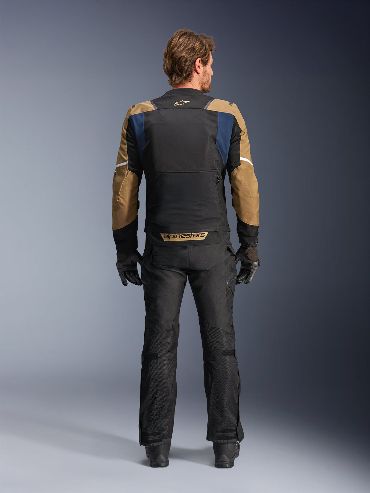 Veste Alpinestars ST-2 Air en bleu profond, kaki foncé et noir, vue arrière sur modèle montrant le large panneau dorsal en mesh et les manches kaki avec rayures blanches, associée à un pantalon technique et des gants noirs pour le touring d'aventure.