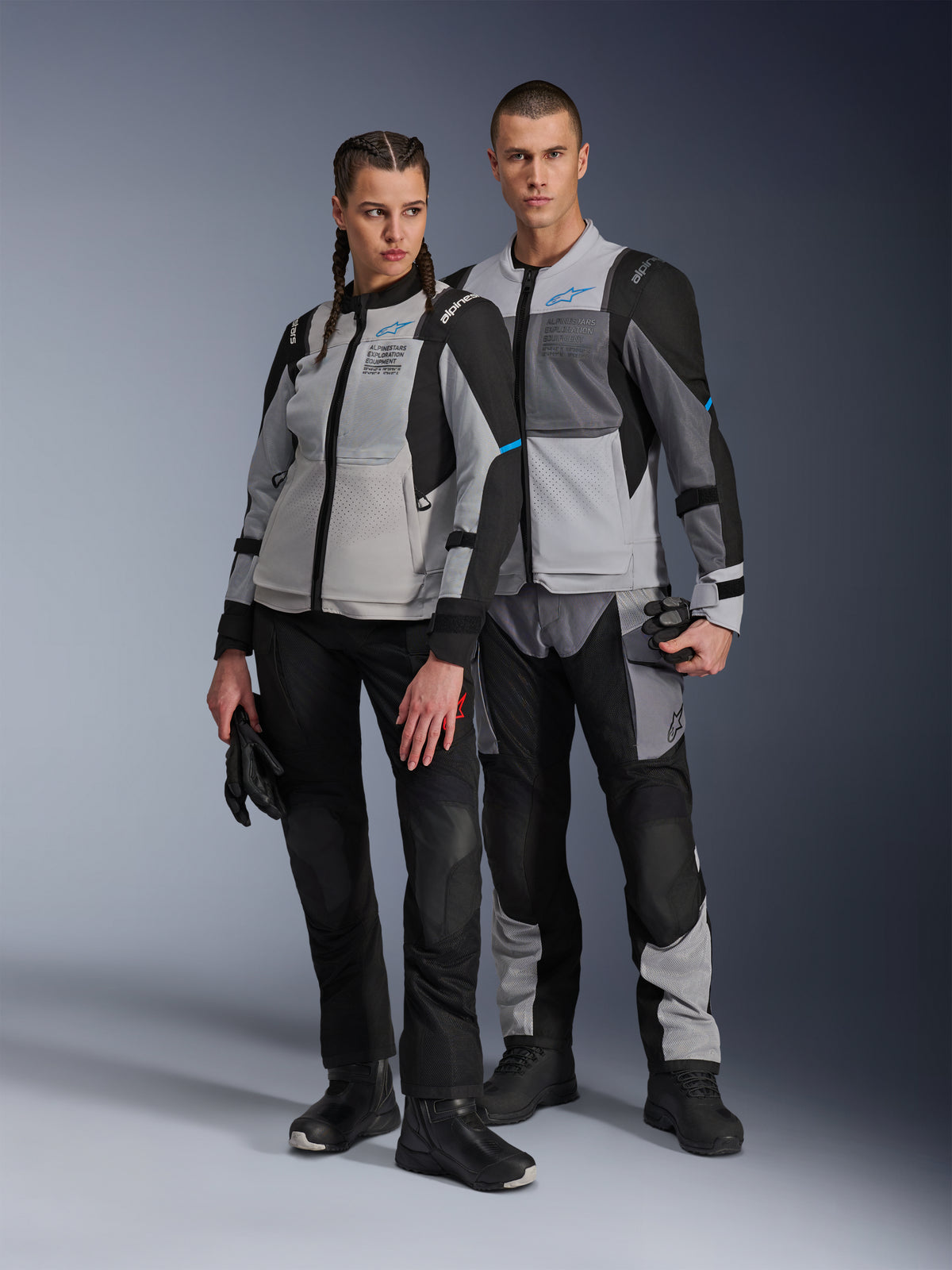 Homme et femme portant des vestes Alpinestars Stella ST-2 Air et pantalons techniques assortis, coloris Gris Glacé et Noir avec accents bleus, dotés de ventilations en mesh et du marquage « ALPINESTARS EXPLORATION EQUIPMENT », photo de couple en studio