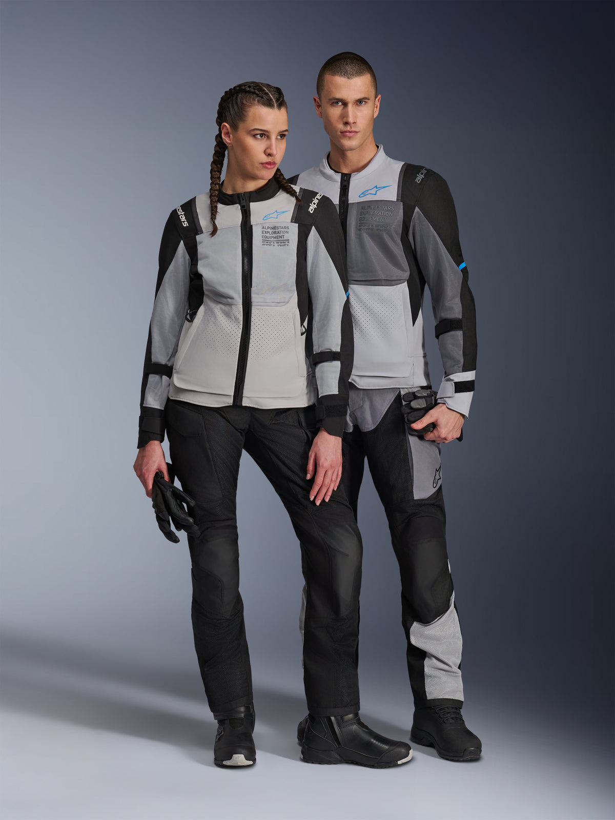 Modèles homme et femme portant les vestes Alpinestars ST-2 Air en Gris Glacé, Gris Foncé et Noir, équipement de moto technique de tourisme estival doté de larges panneaux en mesh et du marquage Exploration Equipment, associé à des pantalons, gants et bottes noirs.