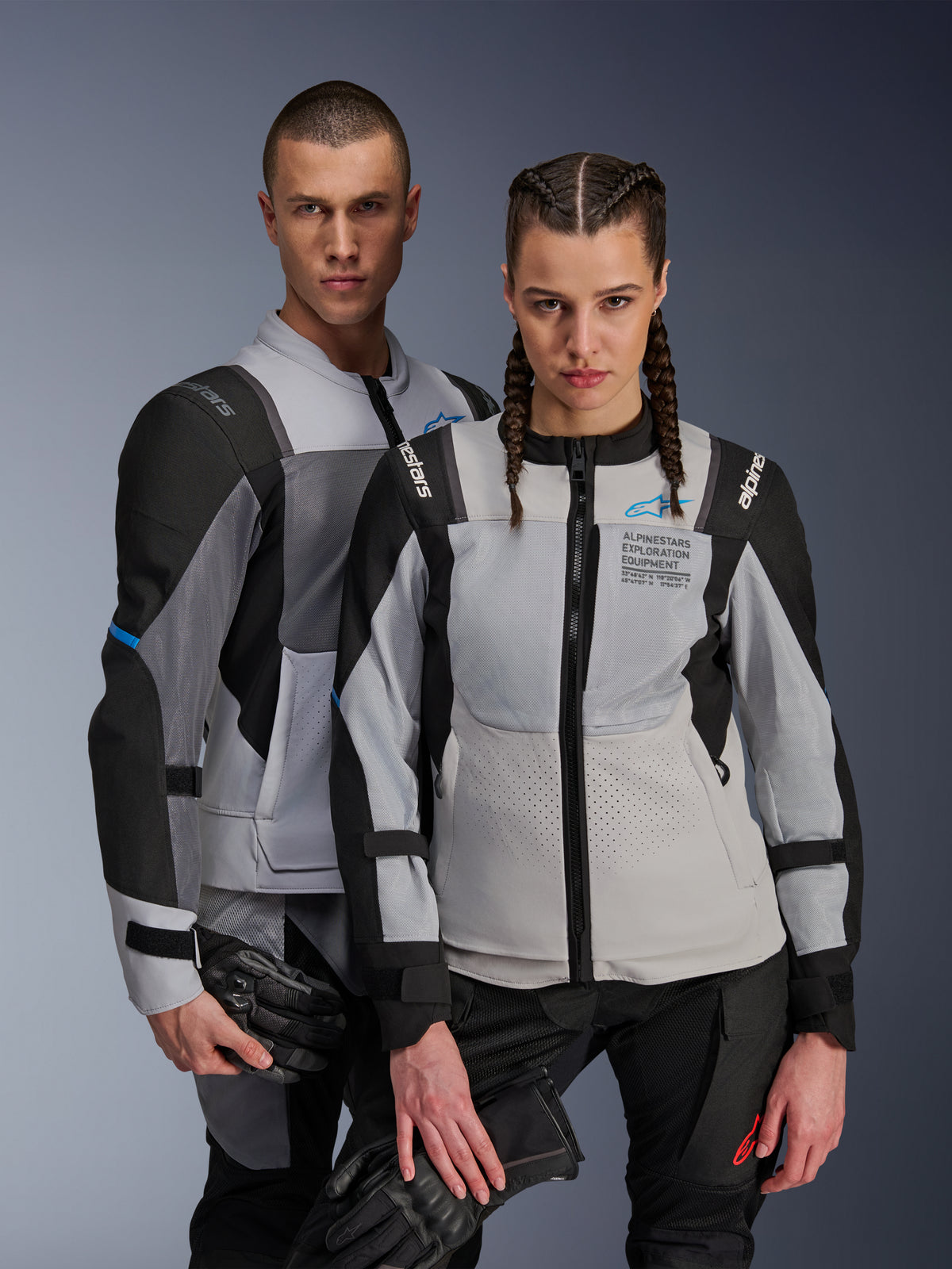 Vestes Alpinestars ST-2 Air et Stella ST-2 Air, gris glacé et noir, vestes de moto en textile avec panneaux en mesh et perforations laser, arborant le marquage « Alpinestars Exploration Equipment », portées par un homme et une femme tenant des gants de moto