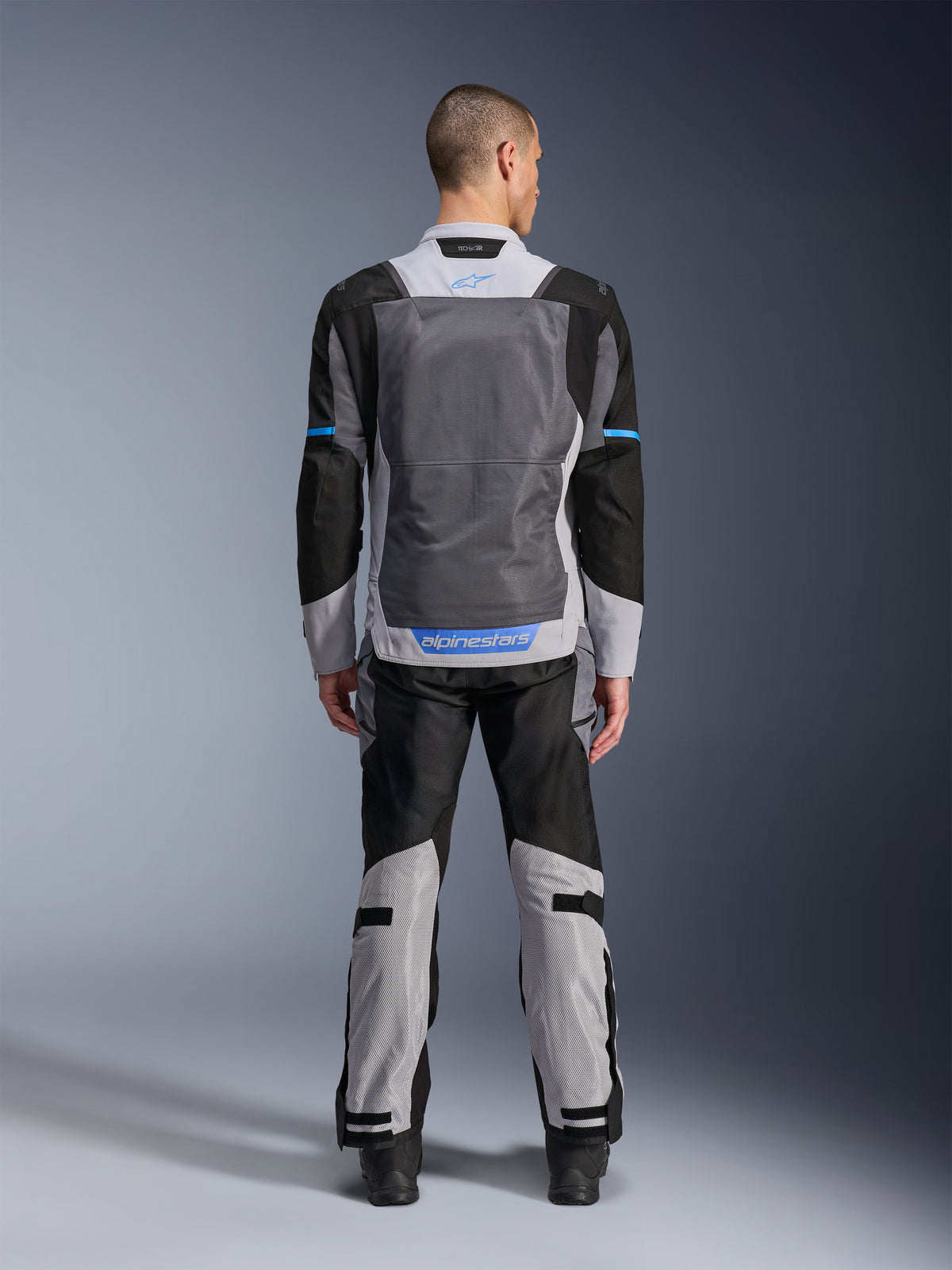 Veste Alpinestars ST-2 Air, Gris Glacé, Gris Foncé et Noir, vue de dos sur mannequin, avec de larges panneaux de ventilation en mesh, des accents bleus et le logo Alpinestars, portée avec un pantalon de moto assorti pour le touring d'aventure