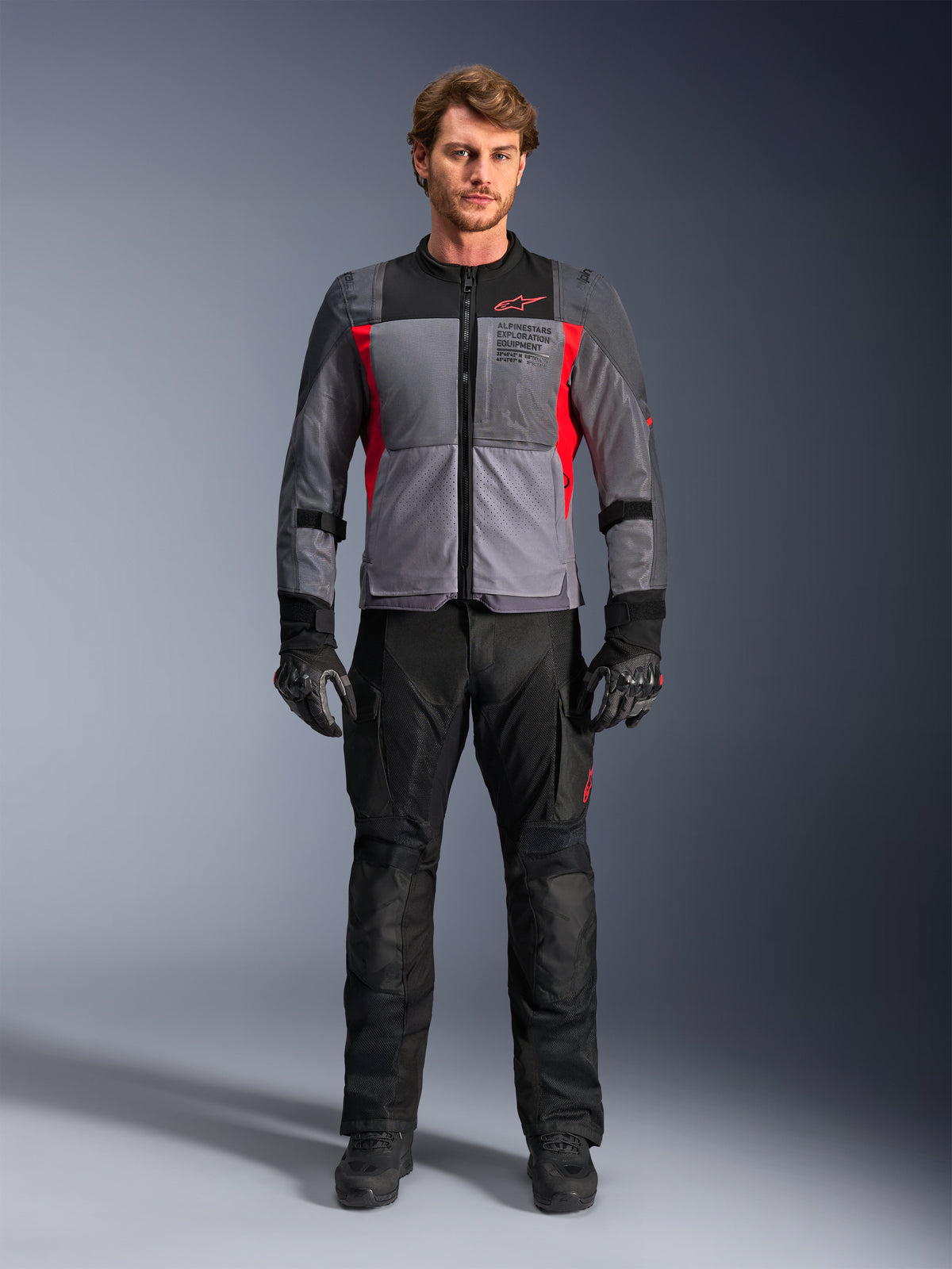 Veste Alpinestars ST-2 Air en gris foncé et rouge vif, portée par un mannequin homme avec un pantalon et des gants de moto noirs, dotée de larges panneaux en maille pour la circulation de l'air et d'un marquage moderne d'équipement d'exploration