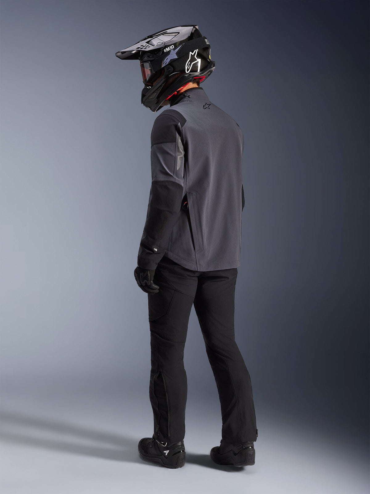 Adventure rider wearing the Maxdura Drystar®XF Laminated Armored Shell in Deep Gray Black, back view. Complete gear setup including black motorcycle pants, a black motocross helmet with Alpinestars branding, and black off-road boots. Technical all-weather riding kit for dual-sport performance.