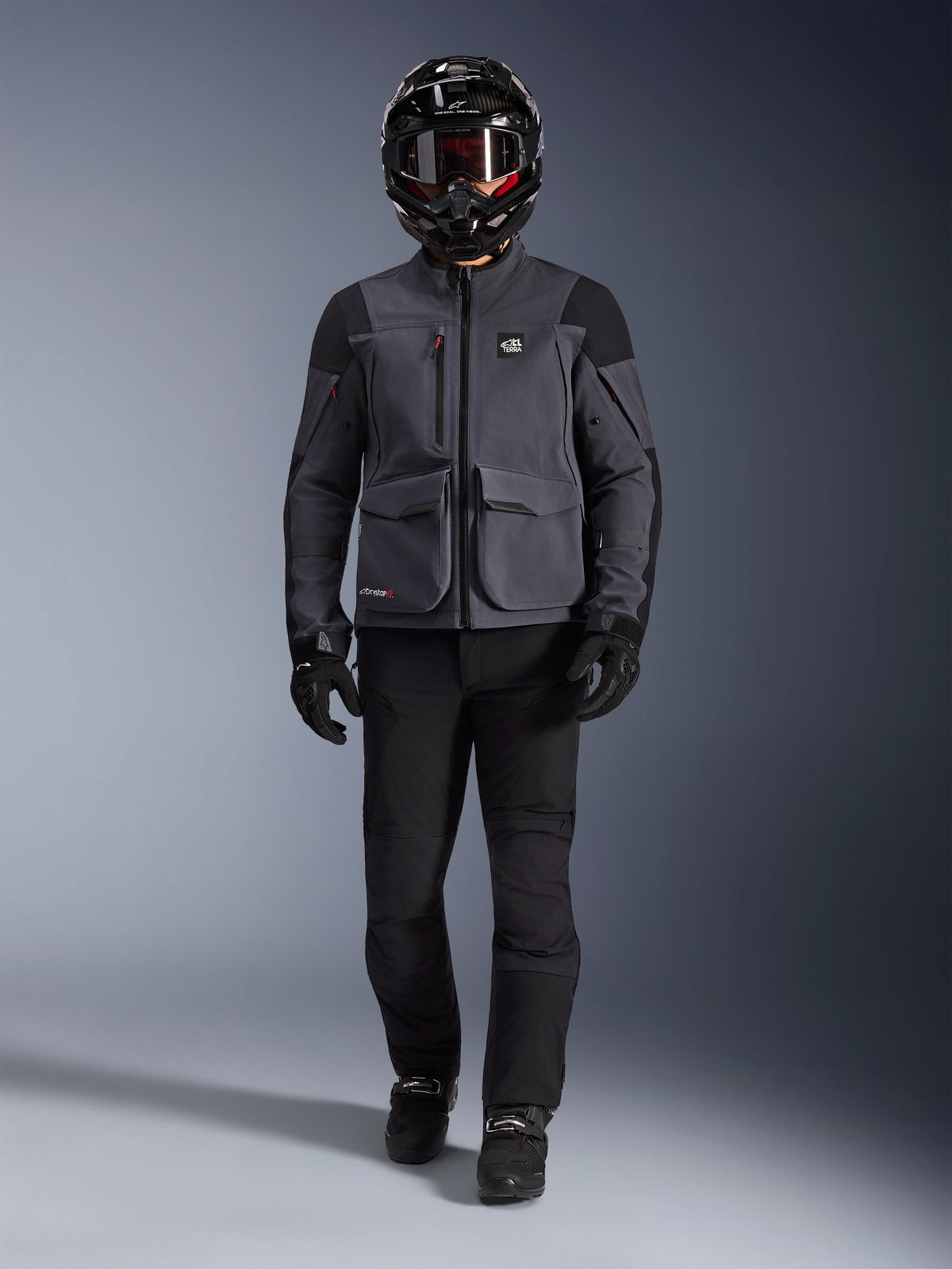 Alpinestars Maxdura Drystar®XF Laminated Armored Shell, adventure motorcycle jacket in Deep Gray and Black, worn with matching black riding pants, a black full-face helmet, gloves, and boots, featuring prominent cargo pockets and ALL TERRA branding for a technical off-road touring look
