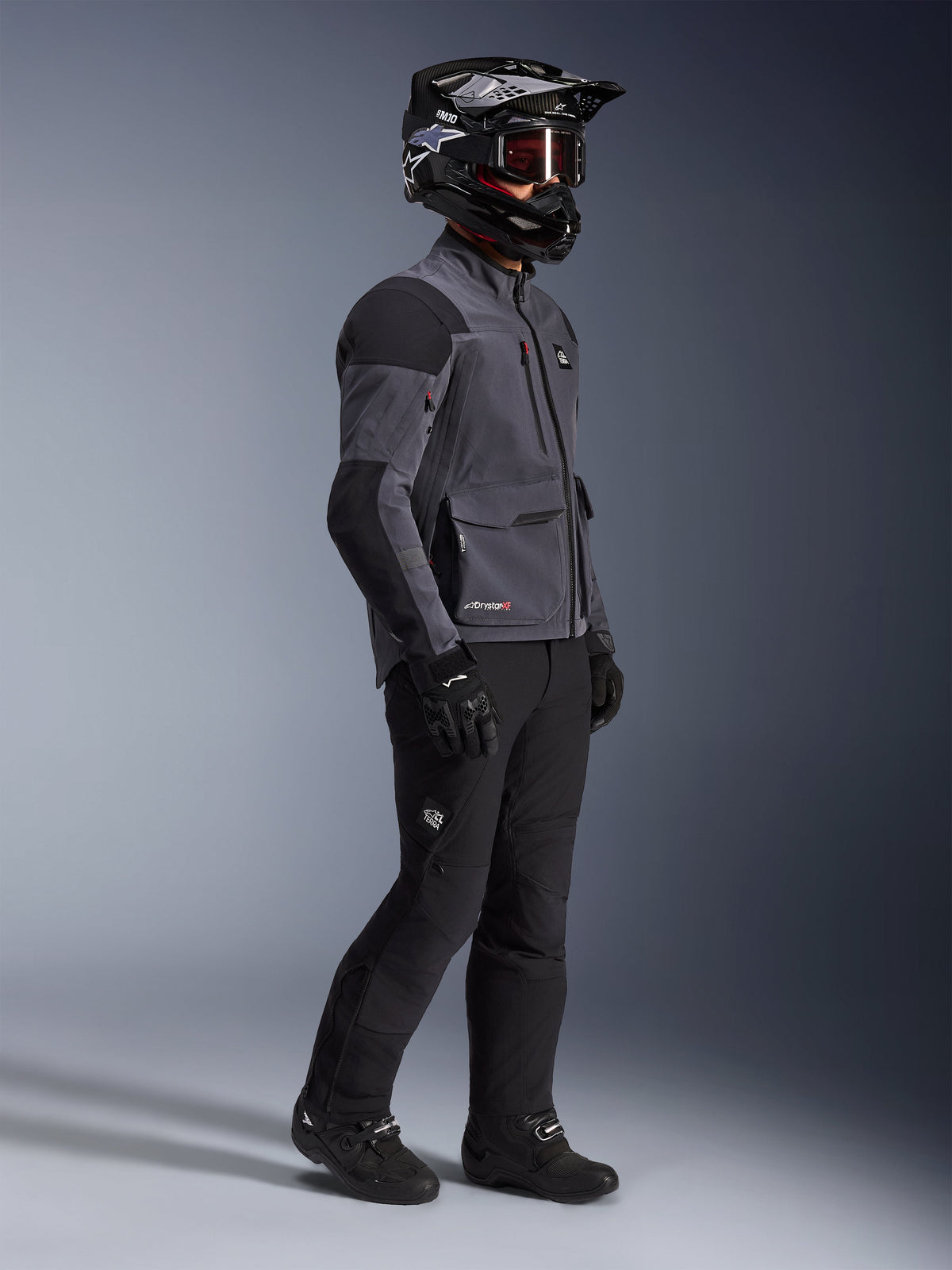 Motorcycle rider wearing Alpinestars Maxdura Drystar®XF Laminated Armored Shell jacket in Deep Gray and Black, paired with black adventure riding pants, black helmet with goggles, black gloves and boots, full-body studio shot highlighting the technical dual-sport gear.