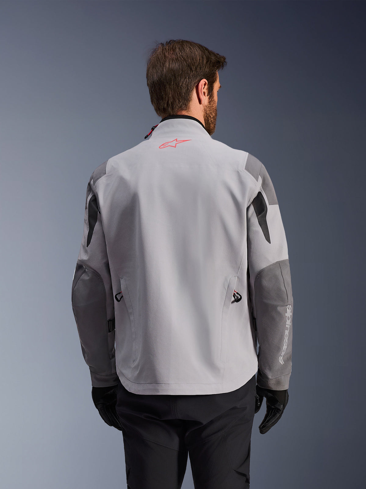 Alpinestars Maxdura Drystar®XF Laminated Armored Shell, vue arrière d'une veste de moto d'aventure, gris moyen et gris foncé, avec un logo étoile rouge sur le col et un marquage vertical blanc sur la manche, portée avec des gants et un pantalon de moto noirs