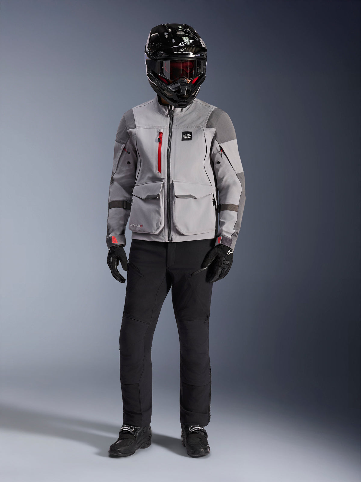 Alpinestars Maxdura Drystar®XF Laminated Armored Shell, veste d'aventure gris moyen et gris foncé avec des accents de fermetures éclair rouges, portée par un pilote avec un casque noir, des lunettes rouges et un pantalon noir, équipement de moto professionnel toutes saisons