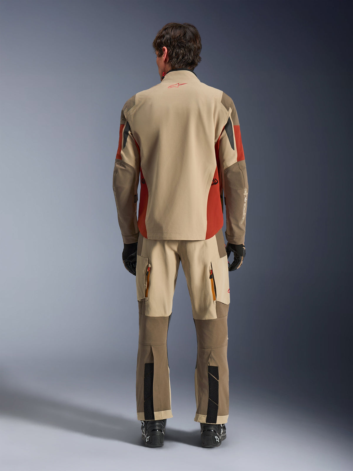 Vue arrière d'un pilote portant la veste Alpinestars Maxdura Drystar®XF Laminated Armored Shell et le pantalon d'aventure assorti, coloris Aluminum Walnut Rust Brown, textile beige et marron avec des accents orange, bottes et gants de moto noirs, équipement de touring professionnel.
