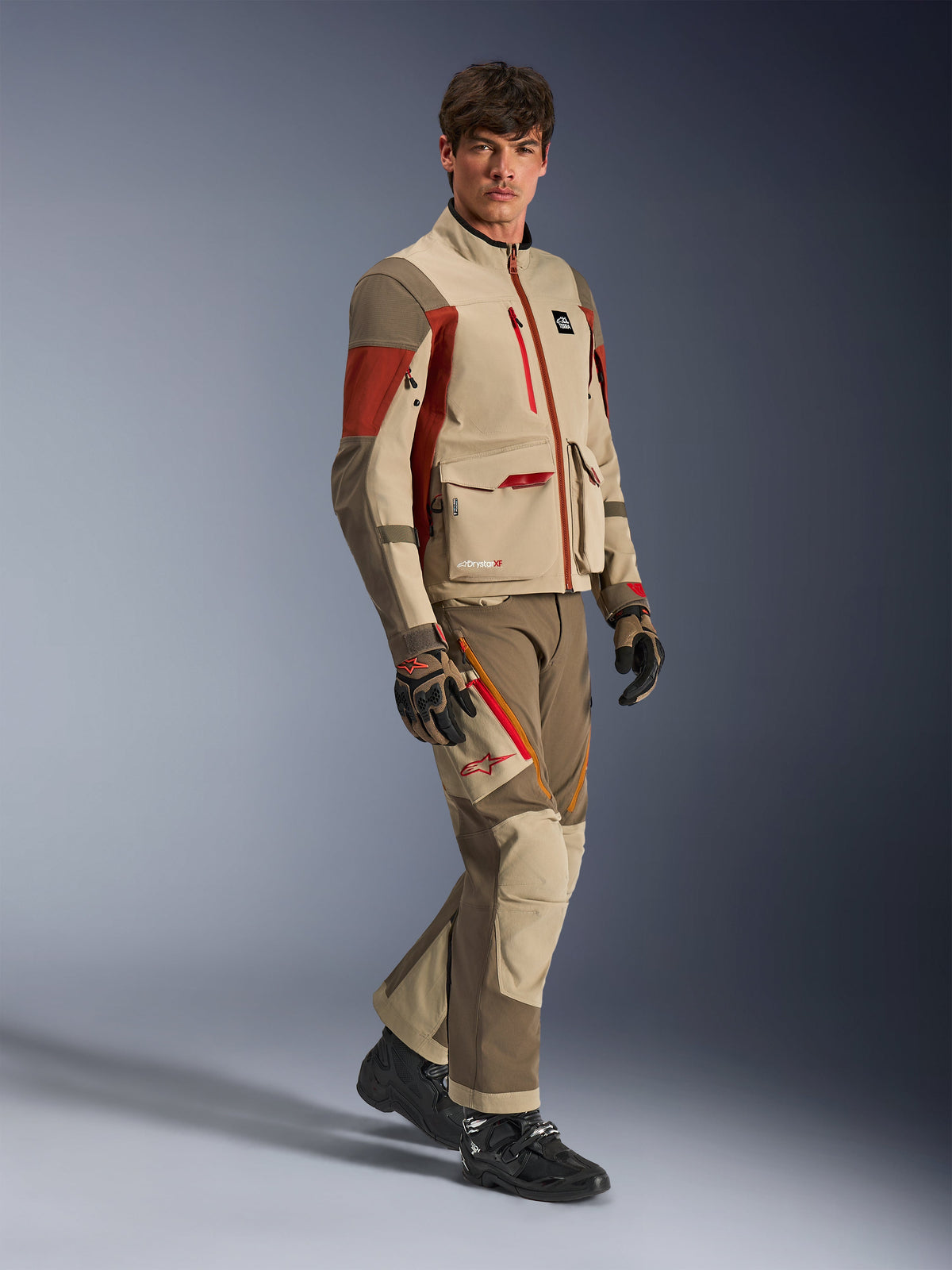 Mannequin homme portant la veste d'aventure Alpinestars Maxdura Drystar®XF Laminated Armored Shell, coloris Aluminum Walnut Rust Brown avec pantalon assorti, gants noirs et marron et bottes noires, dotée d'un design technique multicouche avec poches cargo et détails de zips rouges, tenue coordonnée pour l'aventure touring