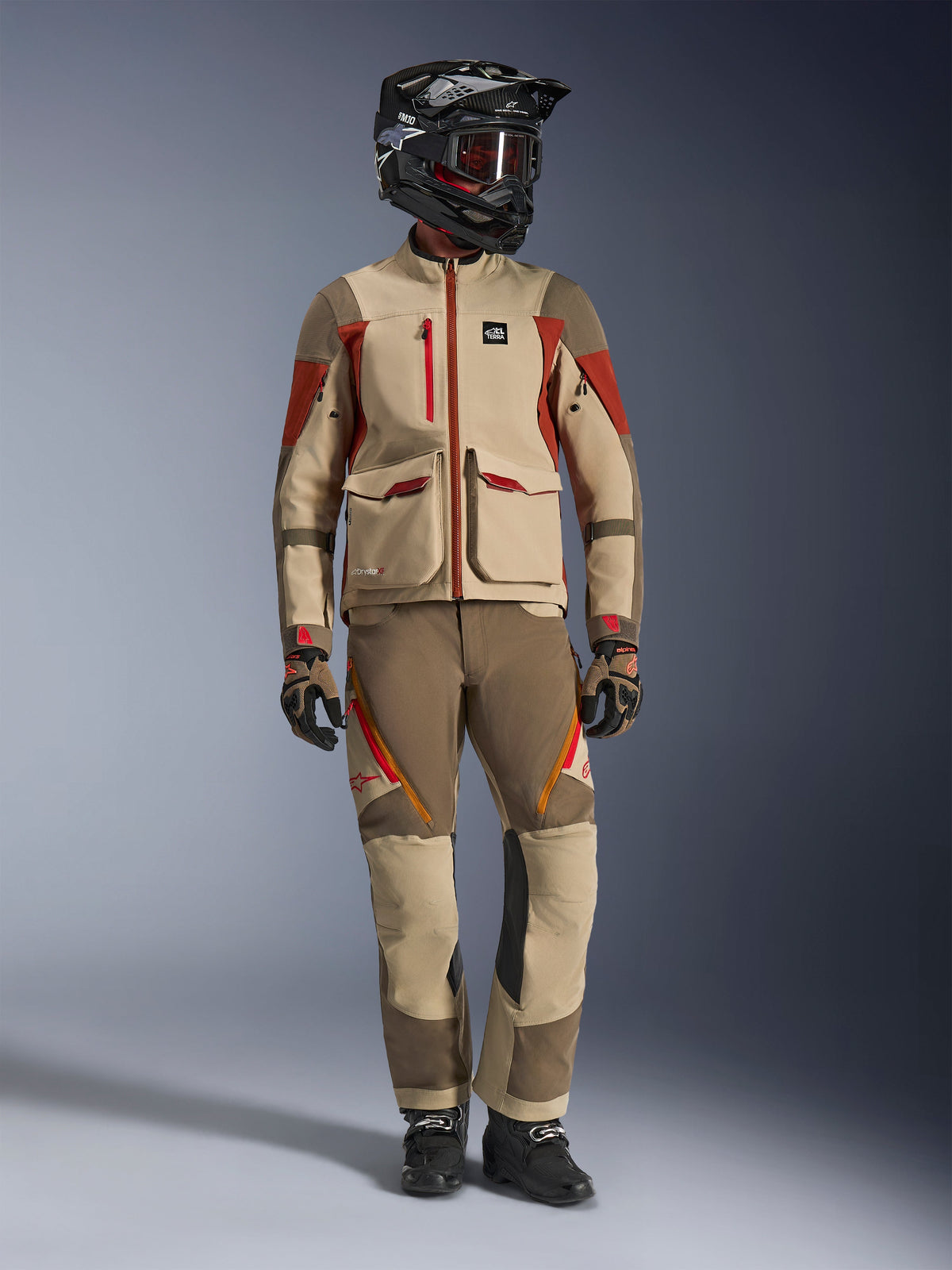 Photo de studio en pied d'un pilote portant la veste Alpinestars Maxdura Drystar®XF Laminated Armored Shell en Aluminum Walnut Rust Brown, assortie d'un pantalon d'aventure, d'un casque tout-terrain noir avec lunettes teintées rouges et de gants techniques, présentant une tenue complète de touring aventure.