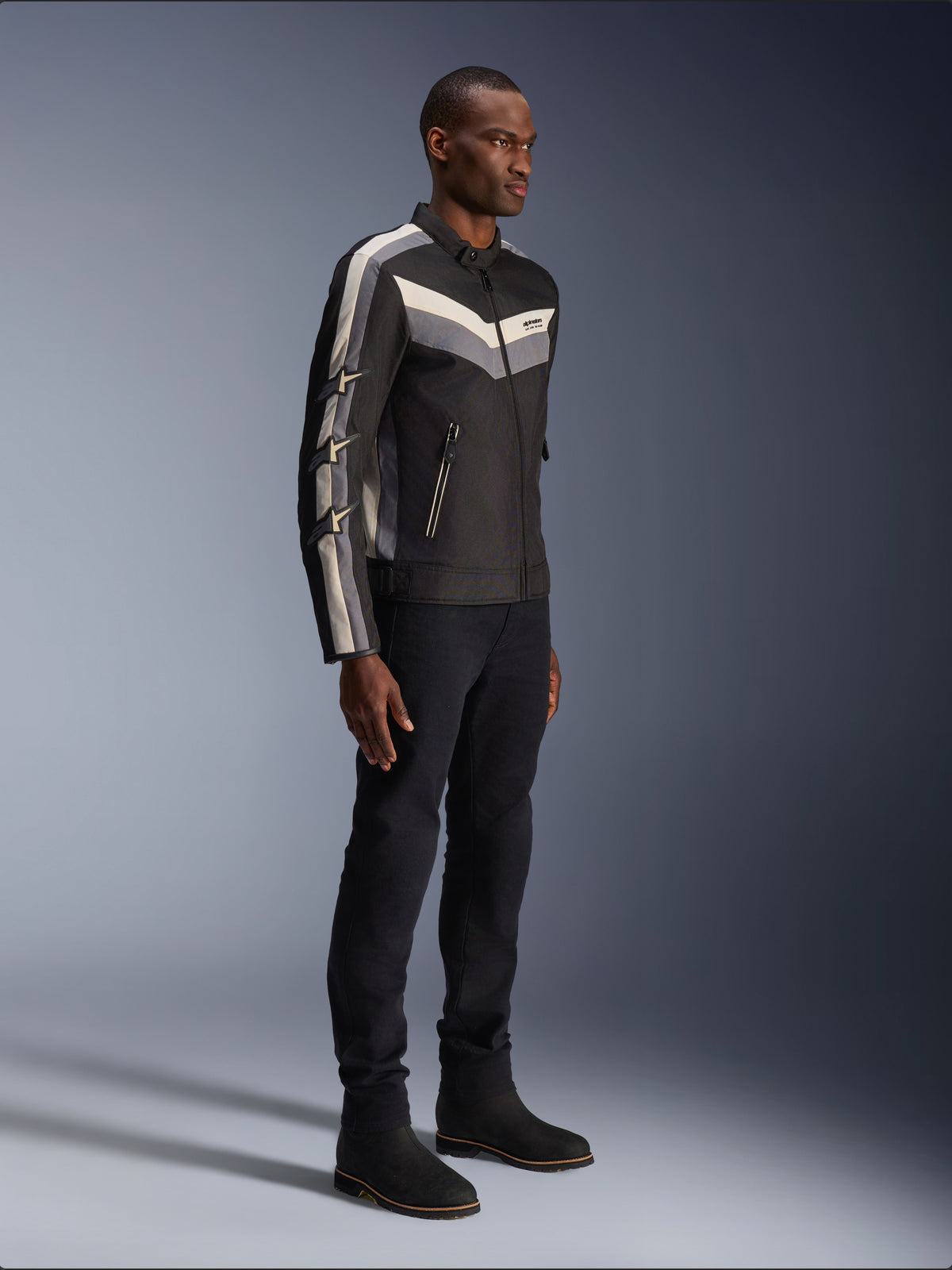 Veste Alpinestars T-Dyno WR, veste de moto en textile, noir avec accents de chevrons gris foncé et écru, coupe biker d'inspiration vintage, portée avec un pantalon et des bottes noirs pour un look routier complet