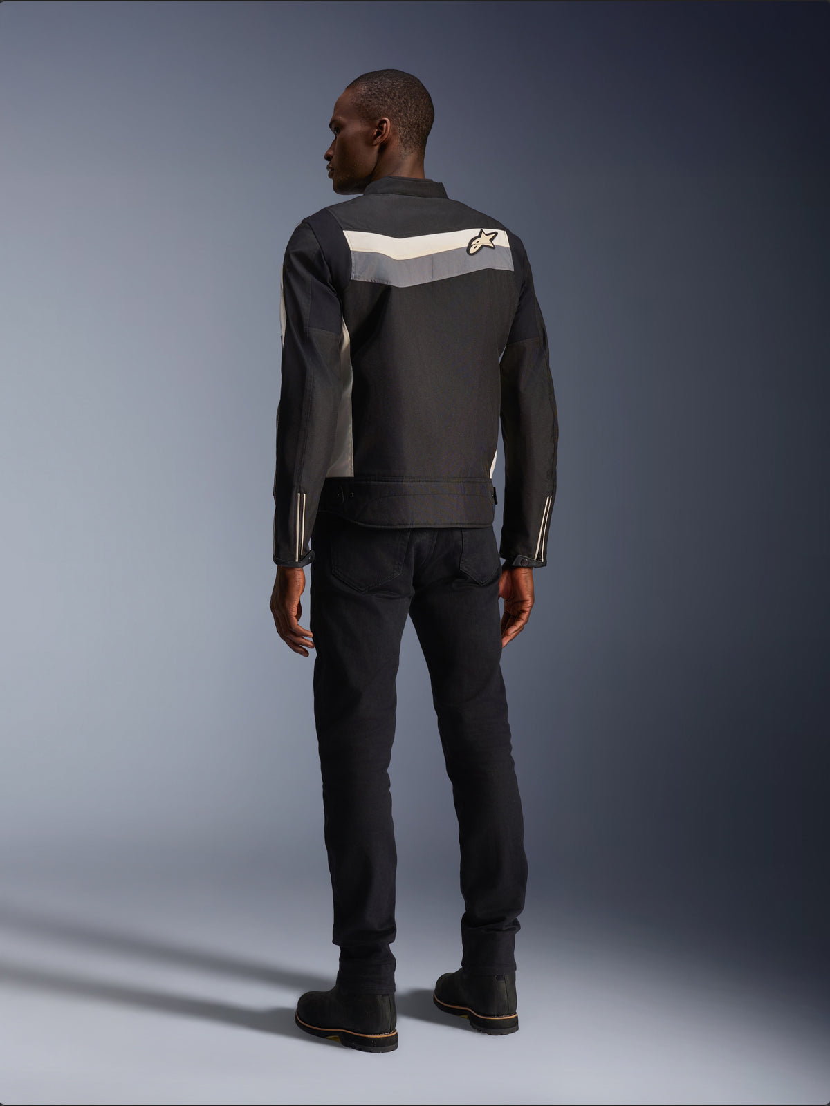 Modèle masculin de dos portant la veste Alpinestars T-Dyno WR en Noir Gris Foncé Écru, veste de moto en textile avec une bande horizontale et le logo étoile au dos, associée à un pantalon de conduite et des bottes noirs, esthétique d'équipement de moto urbain.