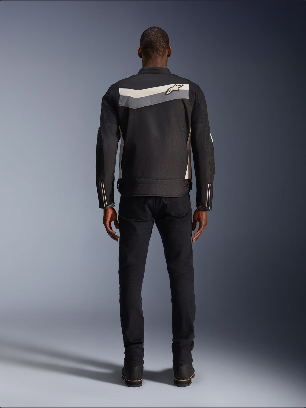 Veste Alpinestars T-Dyno WR, veste de moto en textile, noir, gris foncé et écru, vue de dos montrant une bande horizontale sur les épaules avec un petit logo étoile, portée par un mannequin avec pantalon noir et bottes, équipement de route d'inspiration vintage