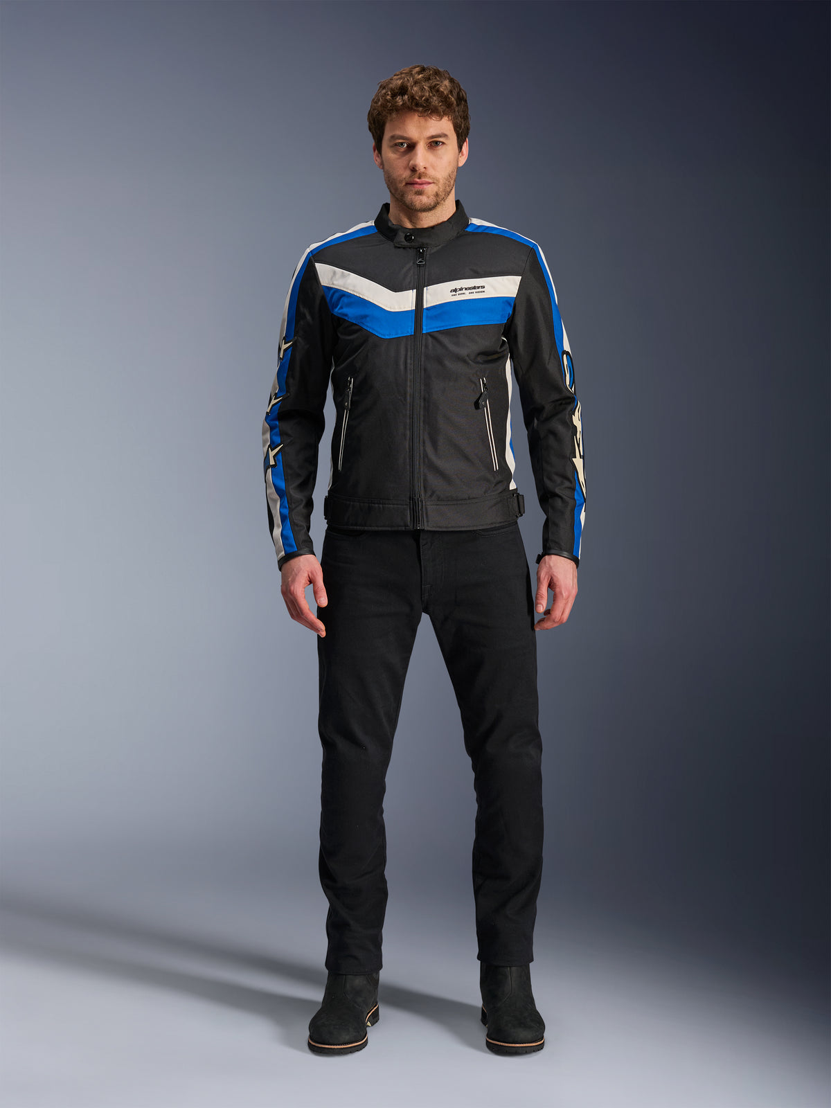 Veste Alpinestars T-Dyno WR, veste de moto en textile, noir, bleu et écru, portée par un mannequin homme avec un pantalon et des bottes noirs, arborant des bandes en chevron sur la poitrine et un design biker héritage