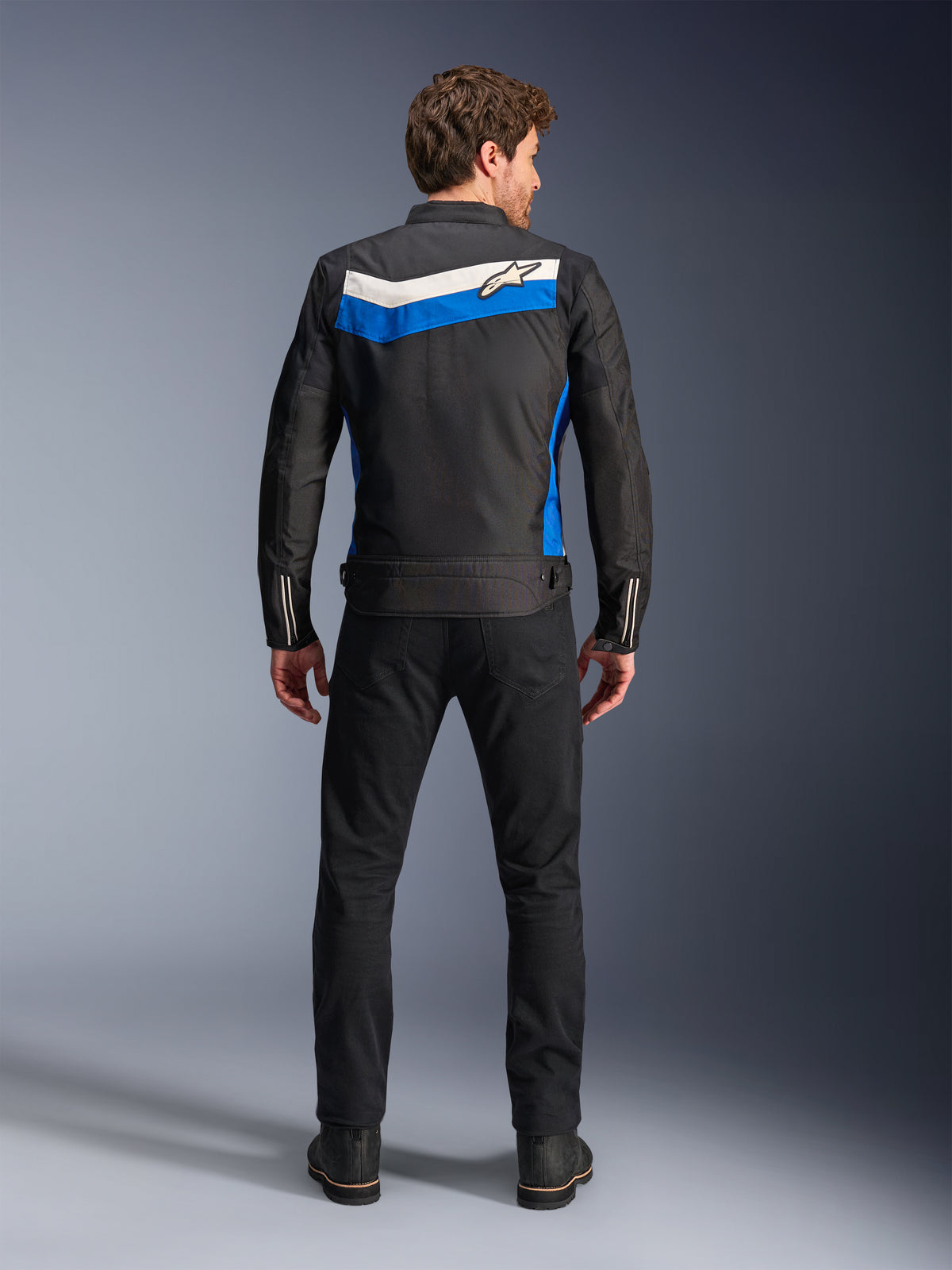 Veste Alpinestars T-Dyno WR, veste de moto en textile, coloris noir, bleu et écru, vue de dos complète sur mannequin, présentant une bande en chevron bleue et blanc cassé avec le logo Alpinestars, associée à un pantalon et des bottes noirs, équipement de conduite urbaine d'inspiration vintage