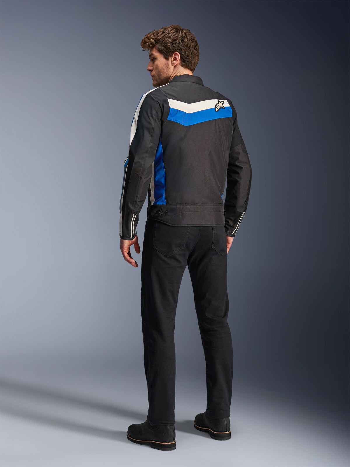 Veste Alpinestars T-Dyno WR en Noir Bleu Écru, vue de dos sur un mannequin homme, avec une bande chevronnée blanche et bleue sur les épaules et un logo étoile, veste de moto en textile d'inspiration vintage, portée avec un pantalon noir et des bottes