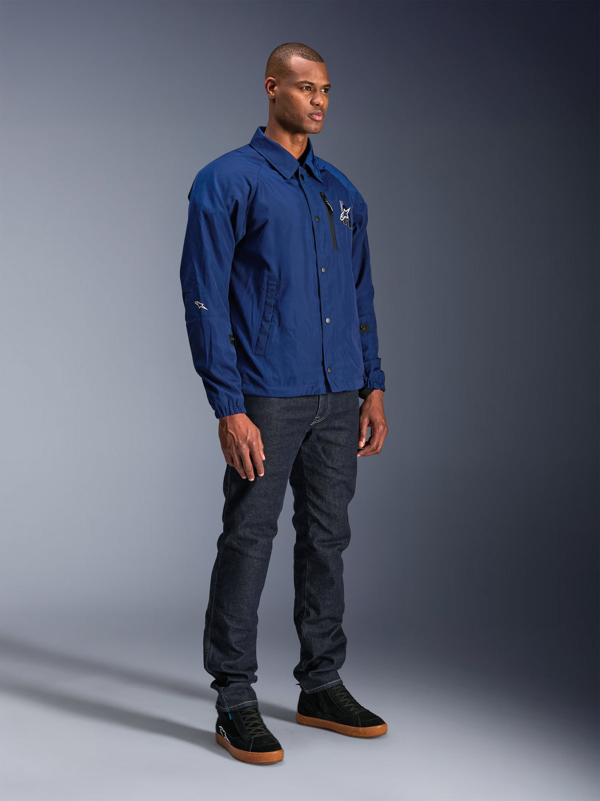 Veste Alpinestars Revved WR, veste de moto bleue, portée par un mannequin homme avec un jean en denim foncé et des baskets noires, présentant une silhouette de veste coach résistante à l'eau avec fermeture à boutons-pression sur le devant et logo minimaliste sur la poitrine.