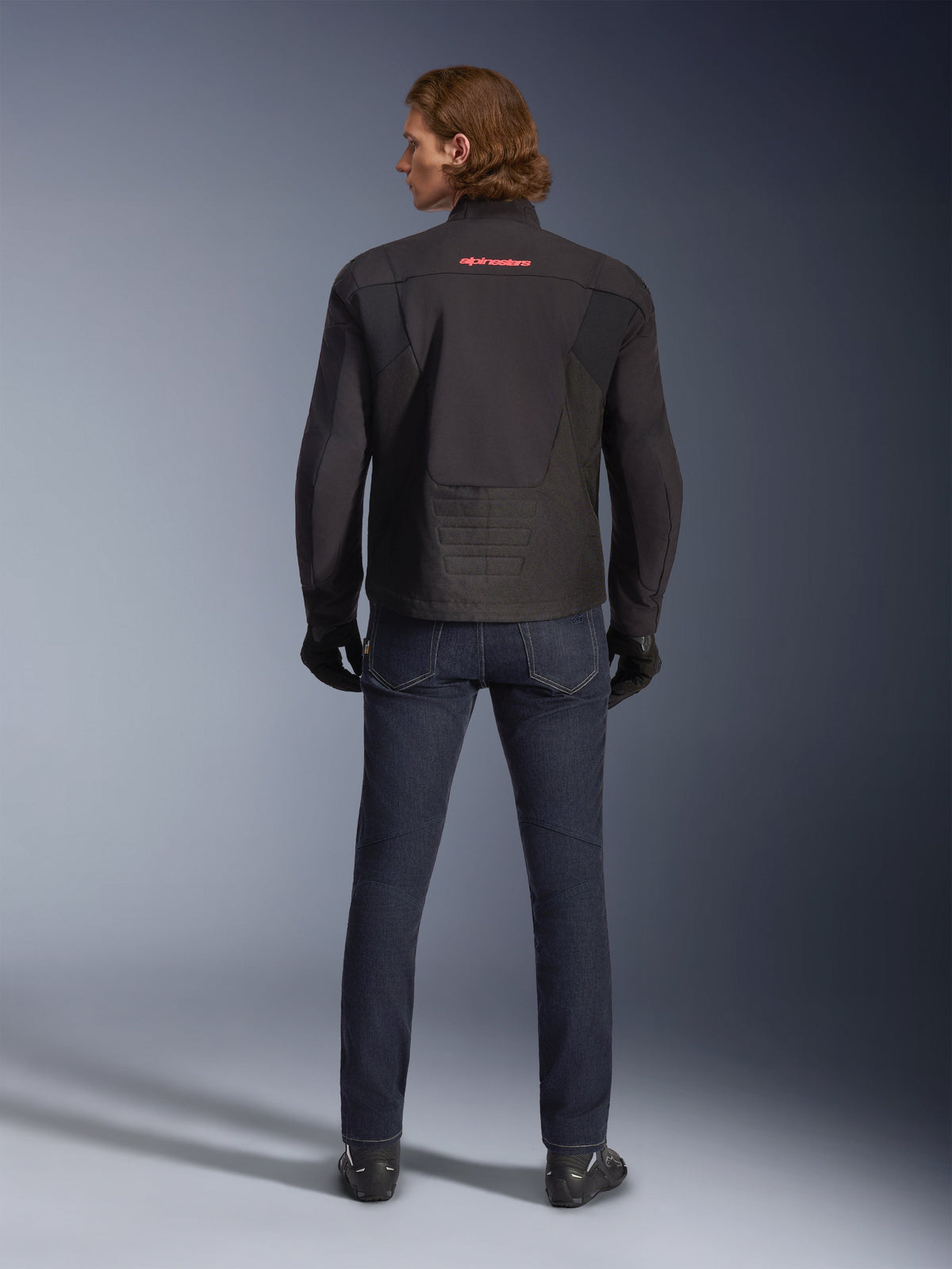 Alpinestars SMX Waterproof Jacket, Black, back view worn by a model, featuring a red logo on the upper back, elongated rear hem for riding coverage, styled with dark blue motorcycle jeans and black gloves.