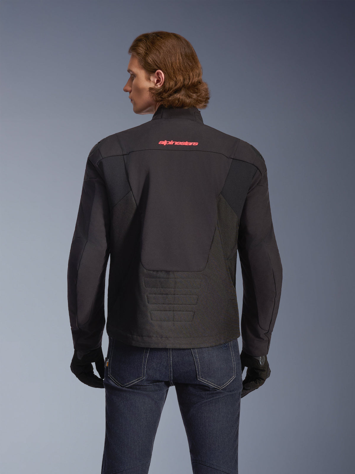 Veste imperméable Alpinestars SMX, noire avec logo rouge vif, vue de dos sur un mannequin, portée avec un jean bleu foncé et des gants noirs, veste de moto en textile pour la route et la conduite sportive