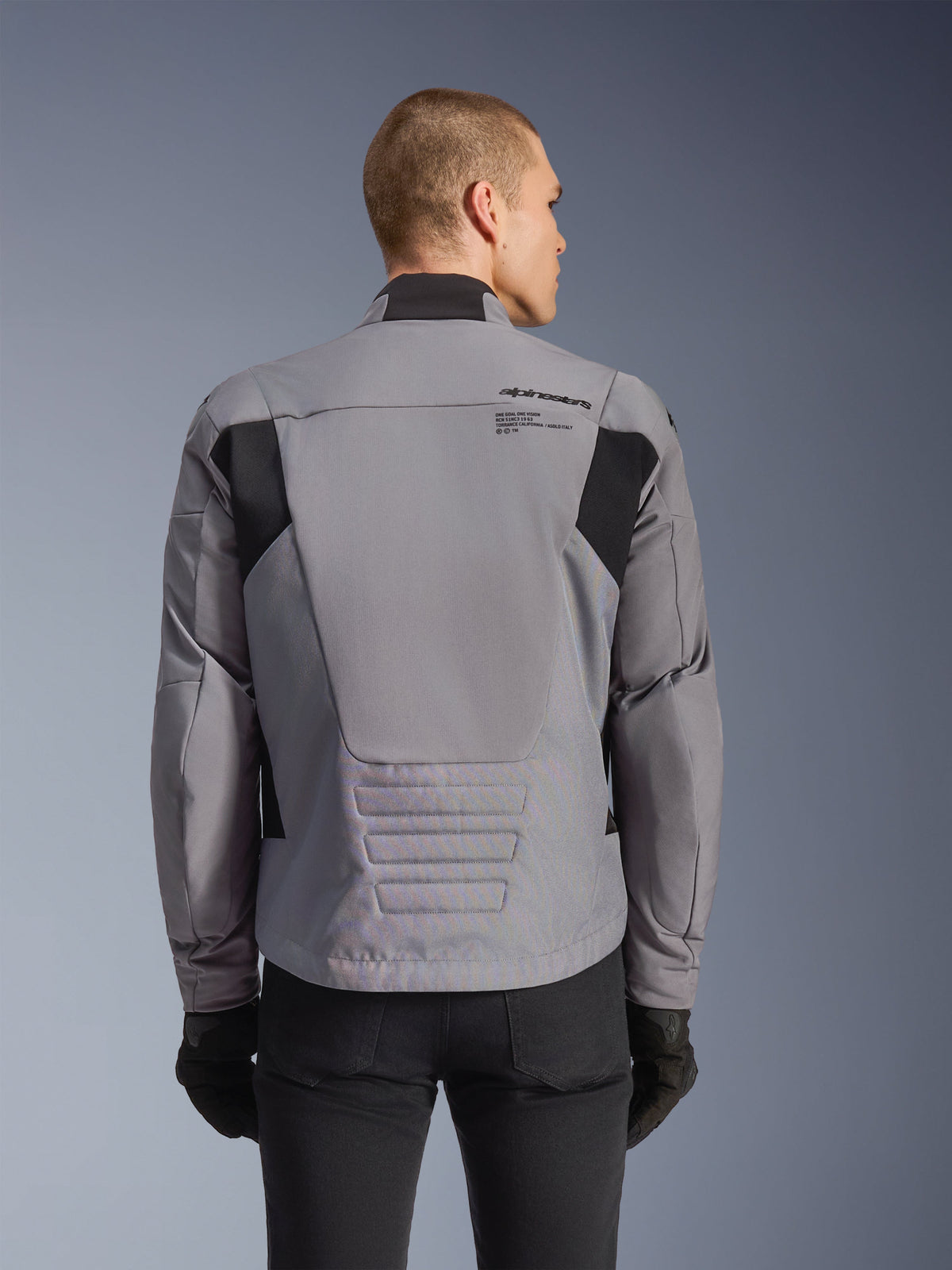 Alpinestars SMX Waterproof Jacket, grise avec accents noirs, vue de dos montrant le marquage sur le haut du dos et le profil arrière allongé, portée par un mannequin avec un pantalon et des gants noirs