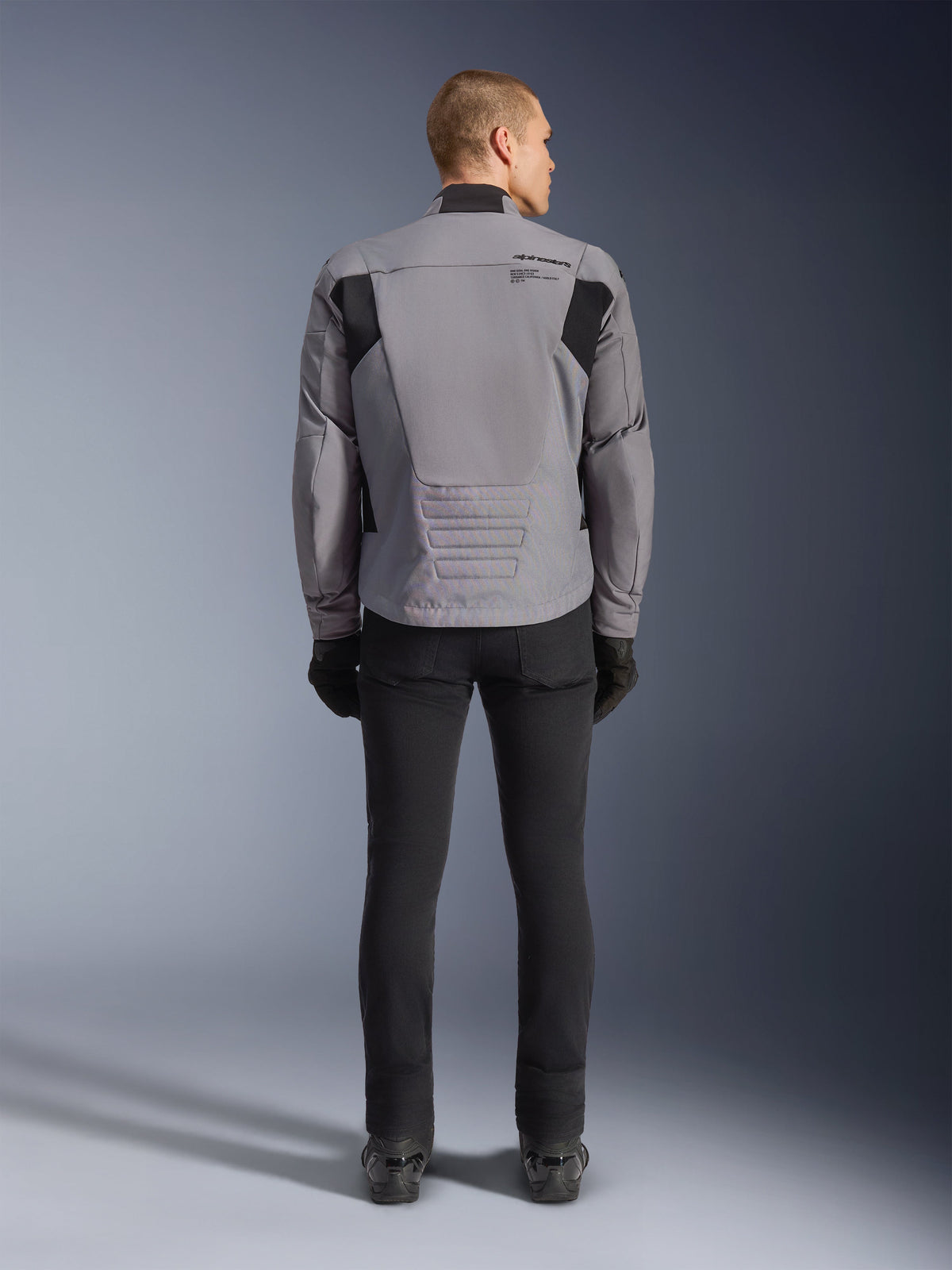 Veste Alpinestars SMX Waterproof, vue arrière, gris foncé et noir, avec empiècements textiles noirs aux épaules et coupe sportive, associée à un pantalon et des gants noirs pour un look moto complet