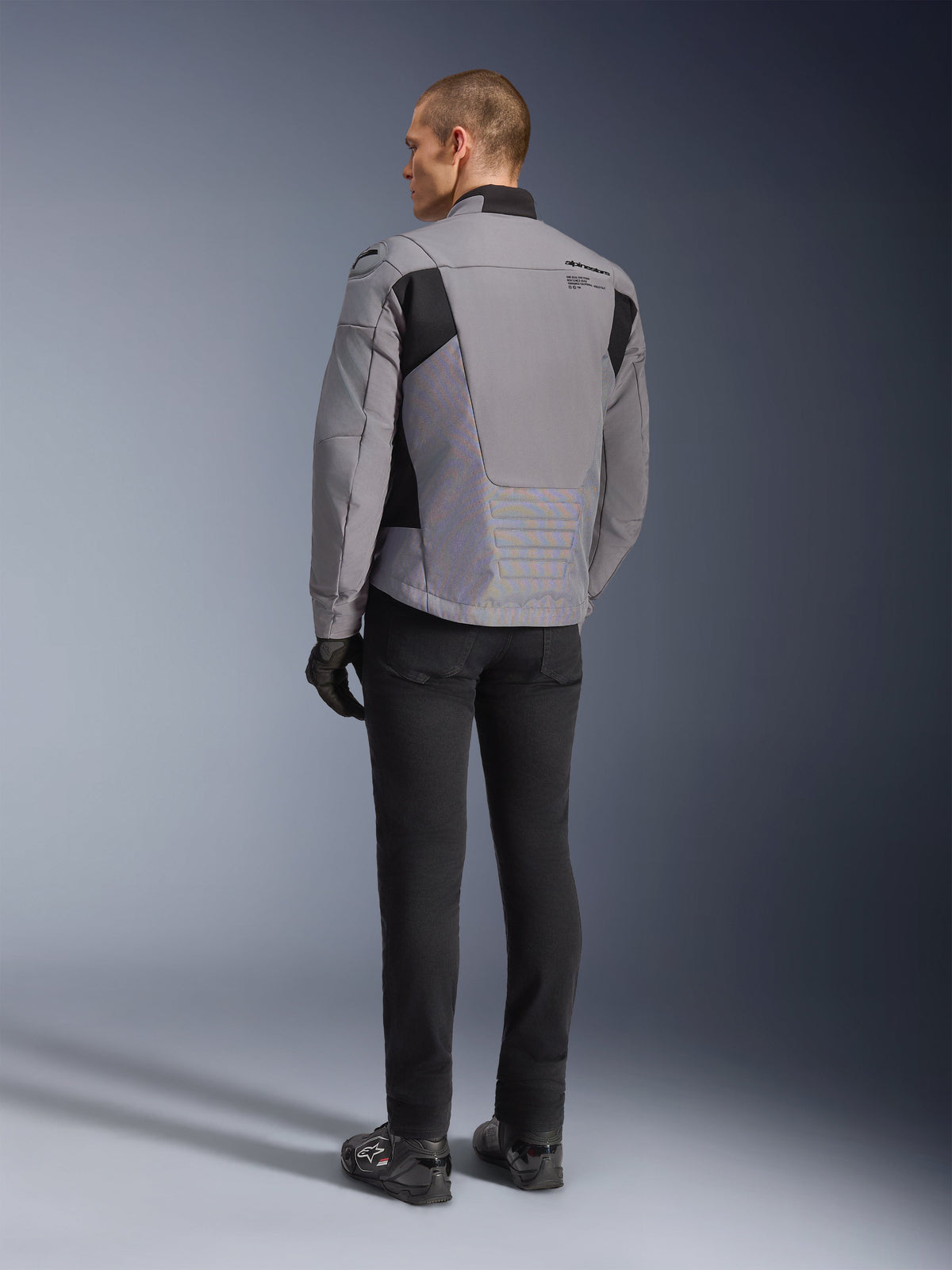 Veste imperméable Alpinestars SMX, gris foncé et noir, vue arrière sur un mannequin, portée avec un pantalon et des bottes noirs, construction en textile technique avec empiècements noirs et logo Alpinestars sur l'épaule.