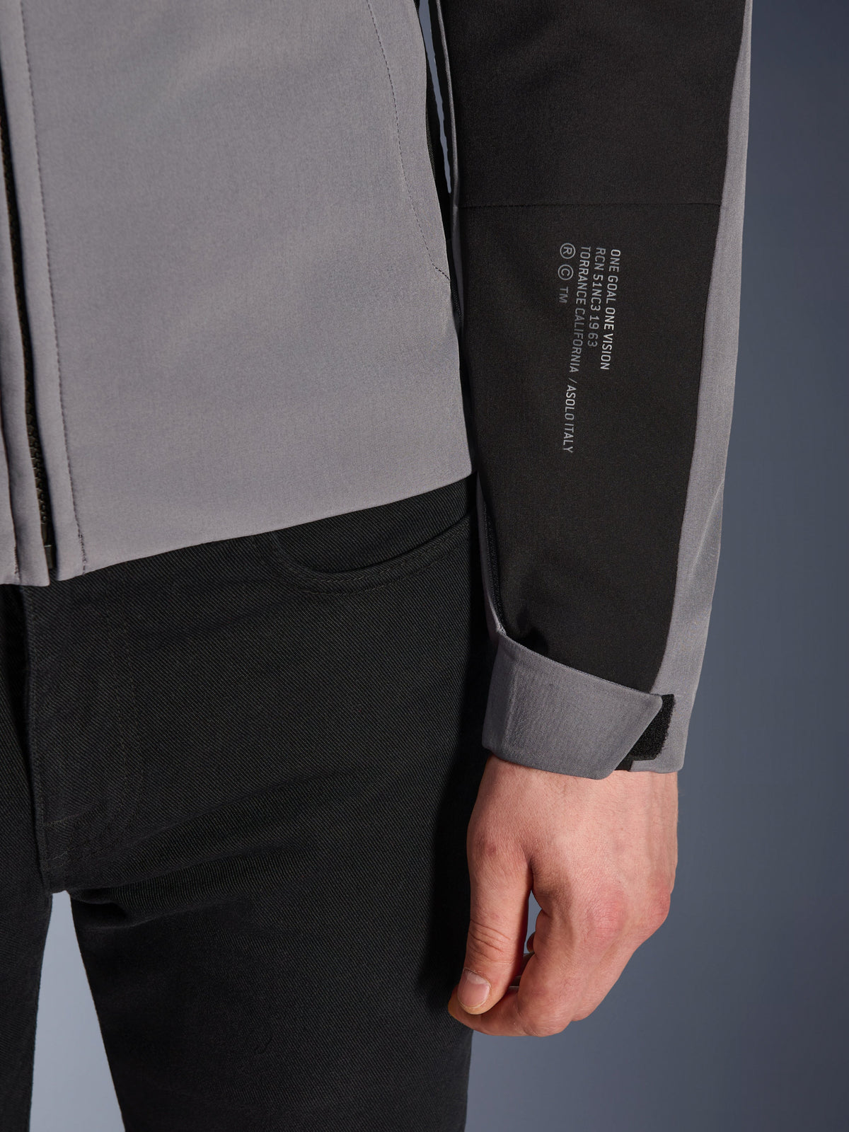 Veste imperméable Alpinestars SMX, gris foncé et noir, gros plan sur la manche avec texte blanc imprimé détaillant l'héritage de la marque, poignet ajustable par Velcro, design textile sportif pour la conduite moto par tous les temps
