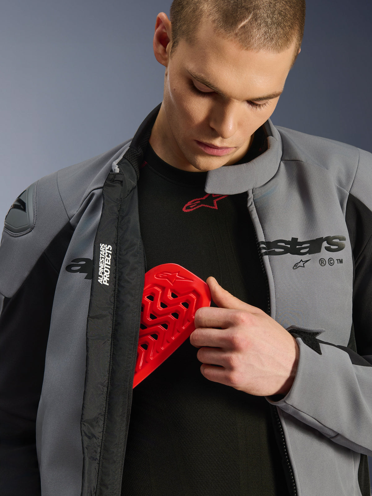 Mannequin portant la veste imperméable Alpinestars SMX en Gris Foncé Noir, insérant une protection de poitrine Nucleon rouge dans une poche interne, illustrant la compatibilité avec les protections et les caractéristiques de sécurité de la veste.