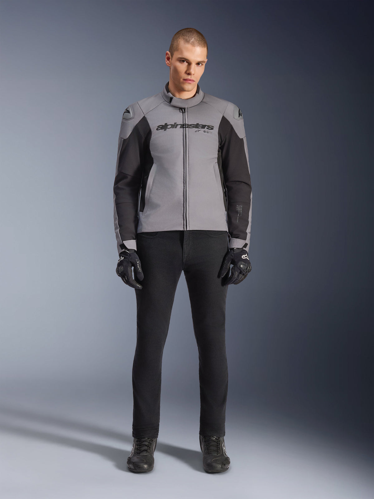 Veste imperméable Alpinestars SMX, gris foncé et noir, vue de face portée par un mannequin homme, veste de moto de sport en textile avec empiècements latéraux noirs et logo Alpinestars sur la poitrine, associée à un pantalon de moto et des gants noirs.