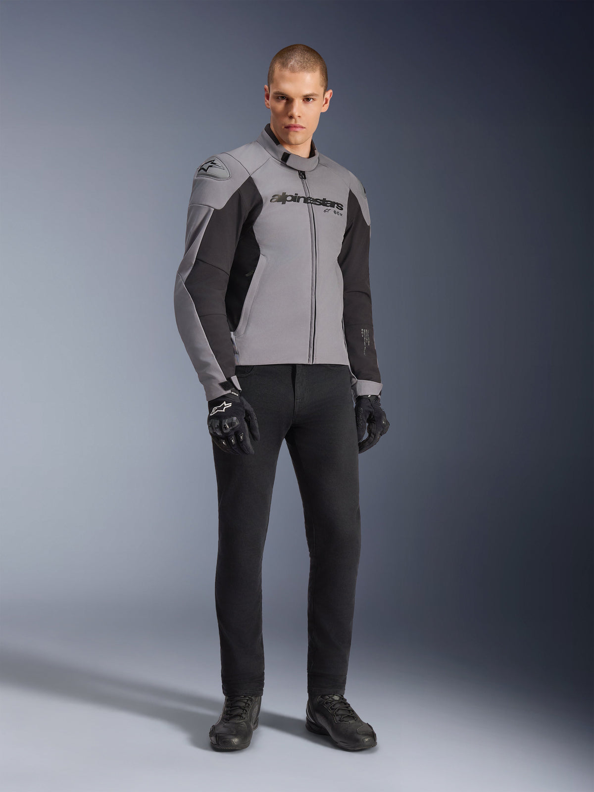 Veste imperméable Alpinestars SMX, veste de moto, gris foncé et noir, portée par un mannequin avec des gants de moto noirs et un pantalon noir, dotée d'un logo imposant sur la poitrine et d'un design technique sport pour la conduite sur route par tous les temps