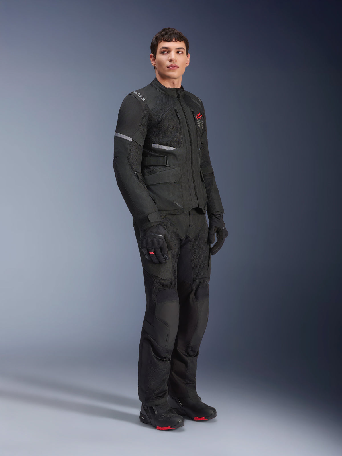 Veste Alpinestars Andes Air Drystar®, noire, portée par un mannequin dans le cadre d'une tenue de moto d'aventure complète avec pantalon, gants et bottes assortis, dotée de panneaux de ventilation en mesh, d'un logo rouge sur la poitrine et de bandes réfléchissantes pour la visibilité.