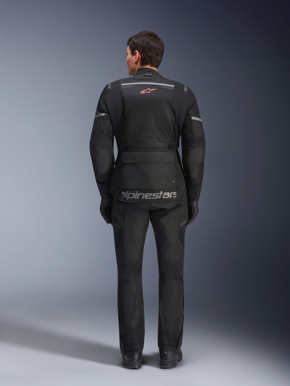 Vue arrière d'un pilote portant la veste Alpinestars Andes Air Drystar® noire, dotée de larges panneaux de ventilation en mesh au dos, d'un logo Alpinestars rouge et de détails réfléchissants, associée à un pantalon et des gants de moto assortis pour le touring aventure.