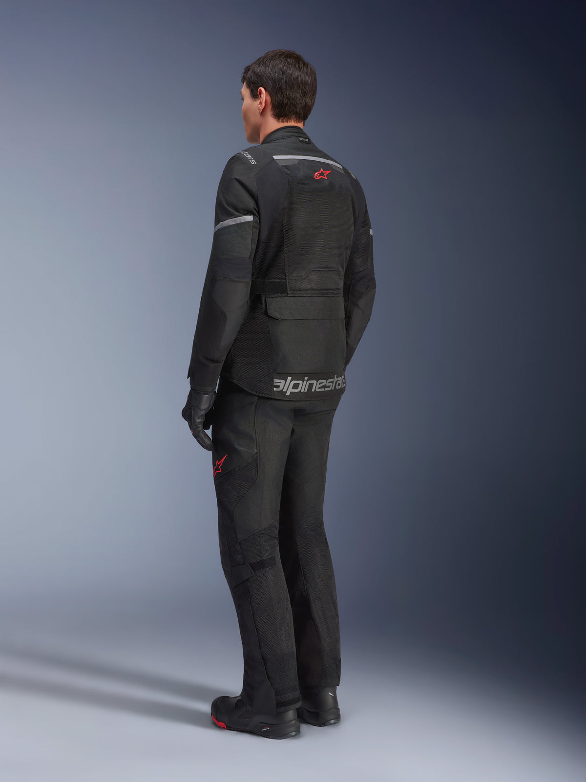 Veste Alpinestars Andes Air Drystar®, Noir, vue arrière sur un motard, dotée de larges panneaux en mesh pour la ventilation, portée avec un pantalon de moto d'aventure et des bottes assortis, mettant en avant le design de touring orienté route et l'image de marque.